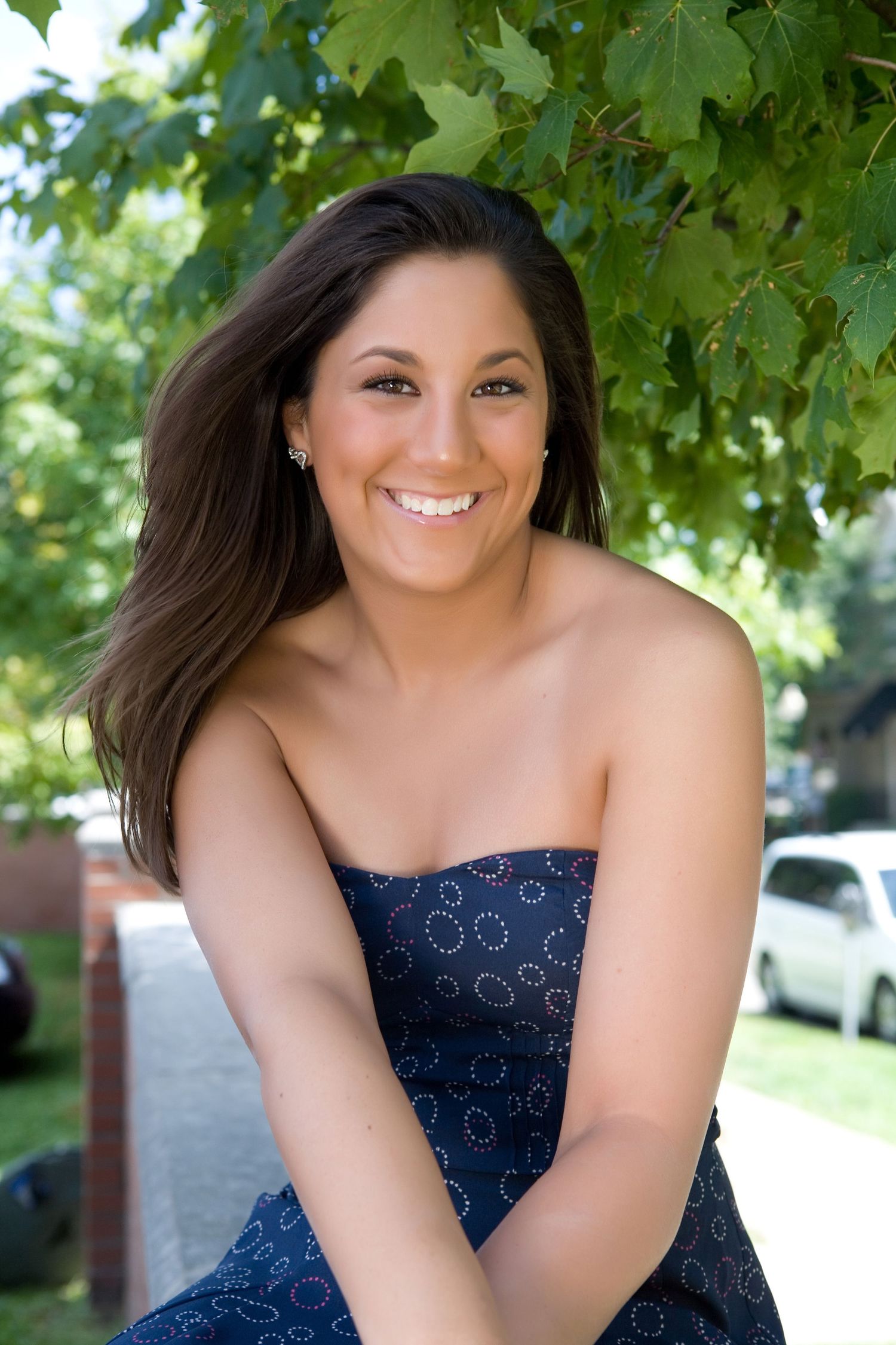 Brunette woman in strapless navy dress smiling outdoors under green tree leaves.