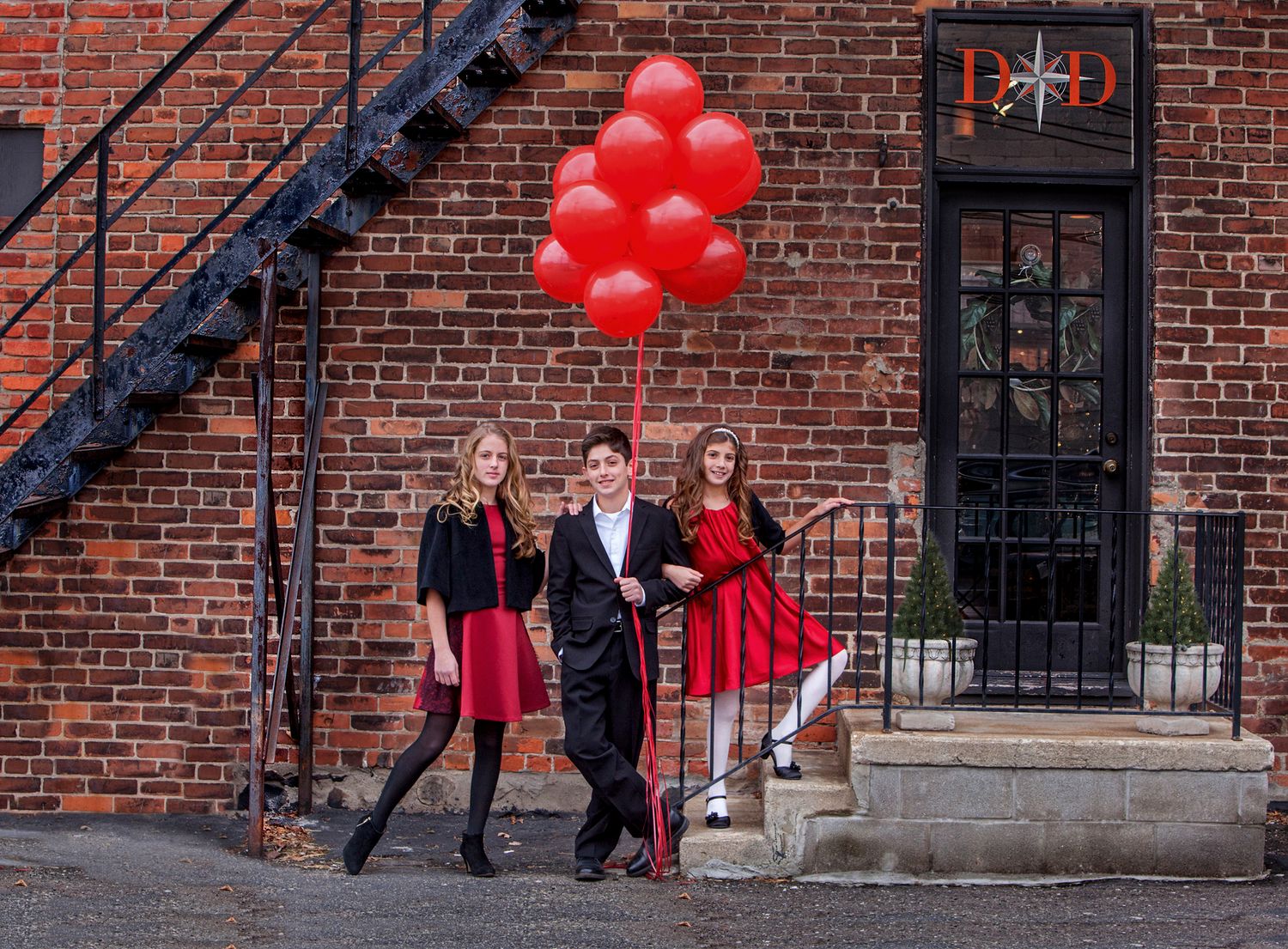 Three teens pose with red balloons outside a brick building, dressed in formal black and red attire.