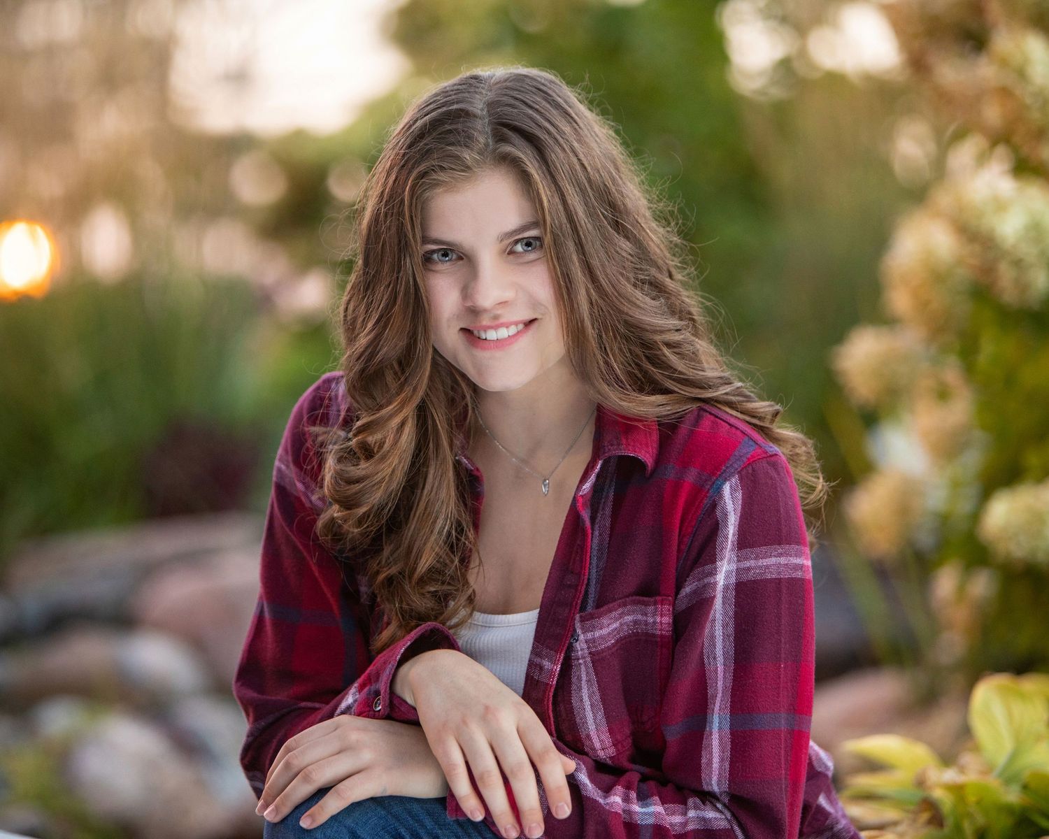 Senior girl smiling outdoors in red plaid shirt among lush garden greenery,by Jocelyn M. Photography in Rochester, MI .