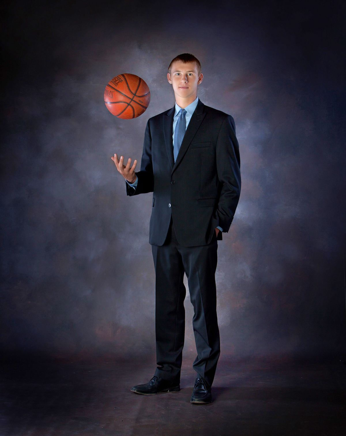 Young man in dark suit balancing a basketball on one hand against a studio backdrop.