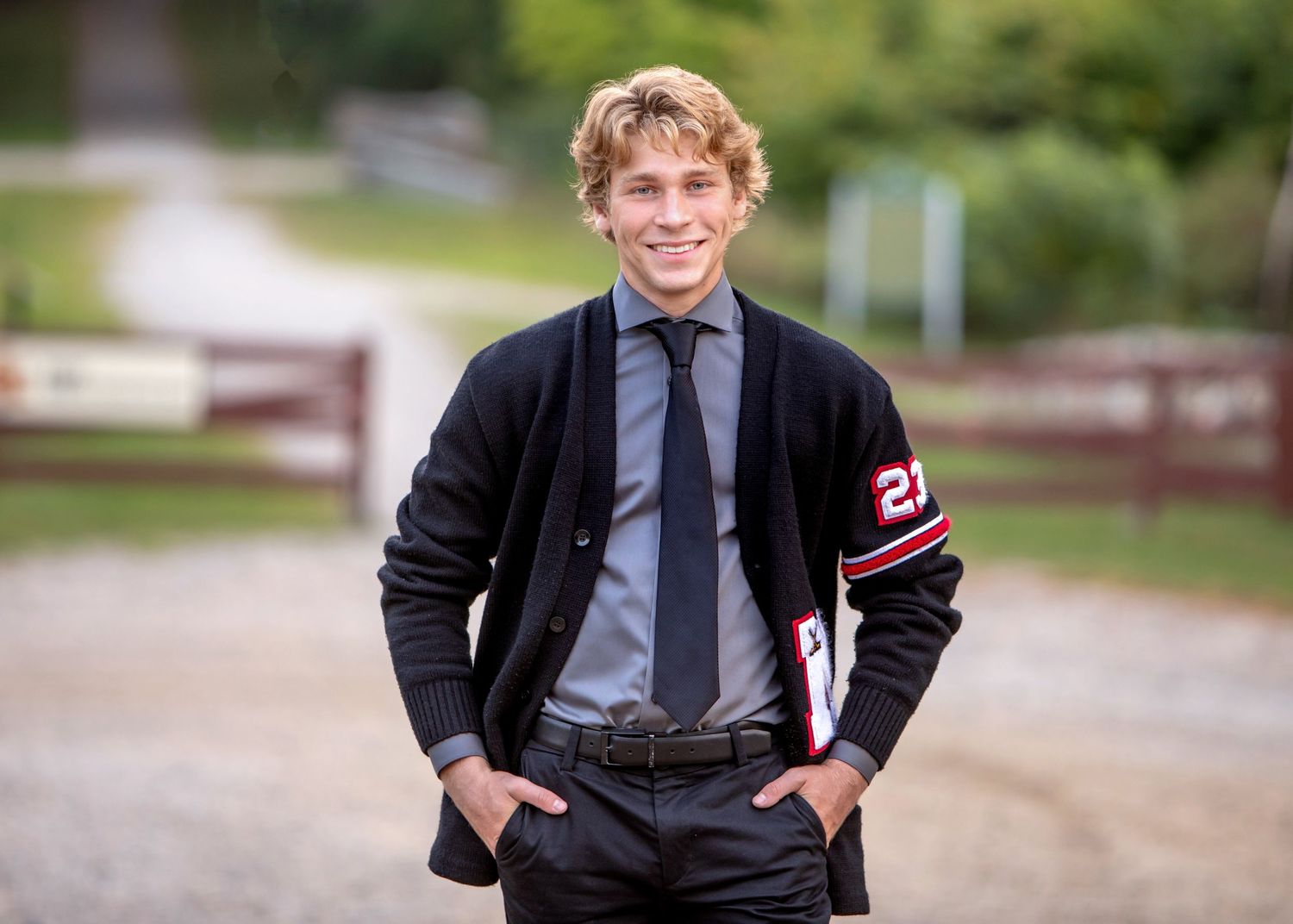Young man in varsity cardigan and tie smiling outdoors near a wooden gate for senior portrait photo. by Jocelyn M. Photography in Rochester,