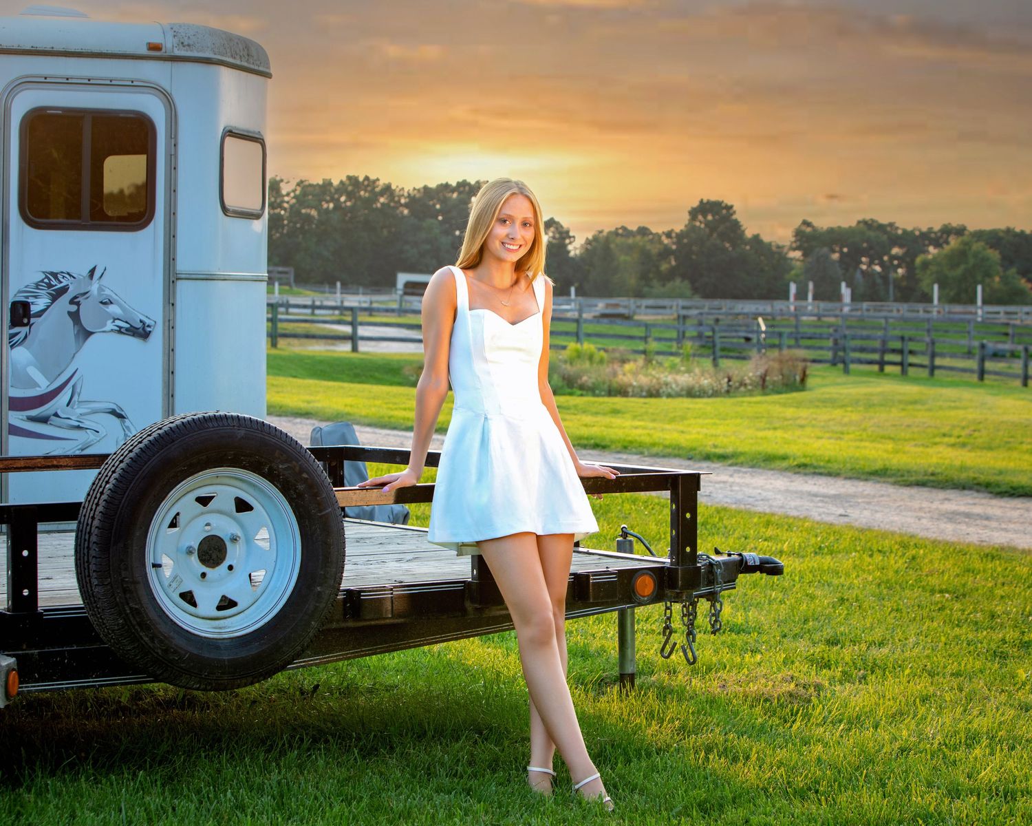 Young woman in white dress posing by a horse trailer at sunset on a scenic farm with wooden fencing.