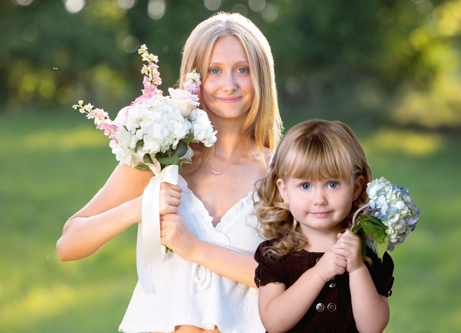 Blonde woman and young girl holding flower bouquets outdoors in a green park setting.