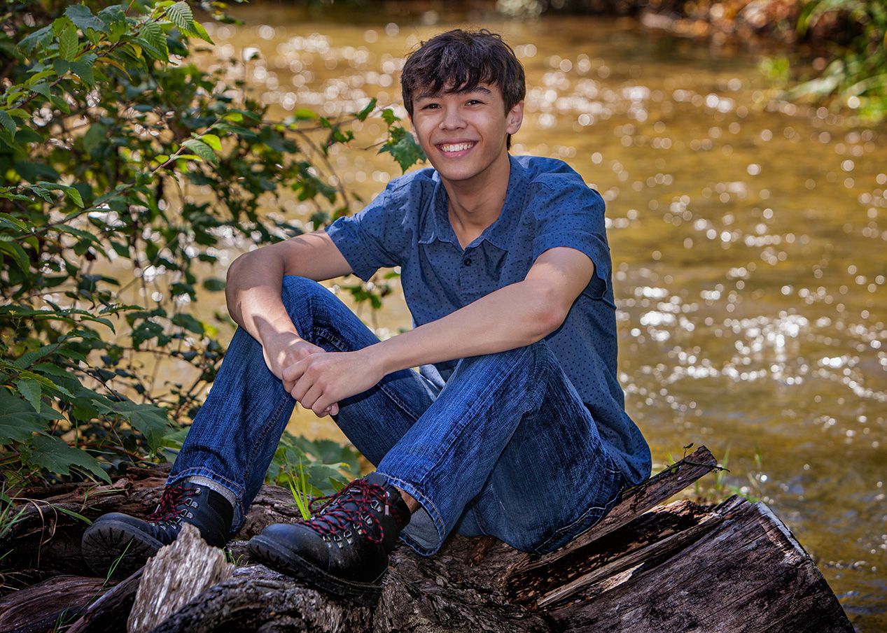 Smiling senior boy sitting on a log by a creek,  surrounded by lush greenery,by Jocelyn M. Photography in Rochester, MI