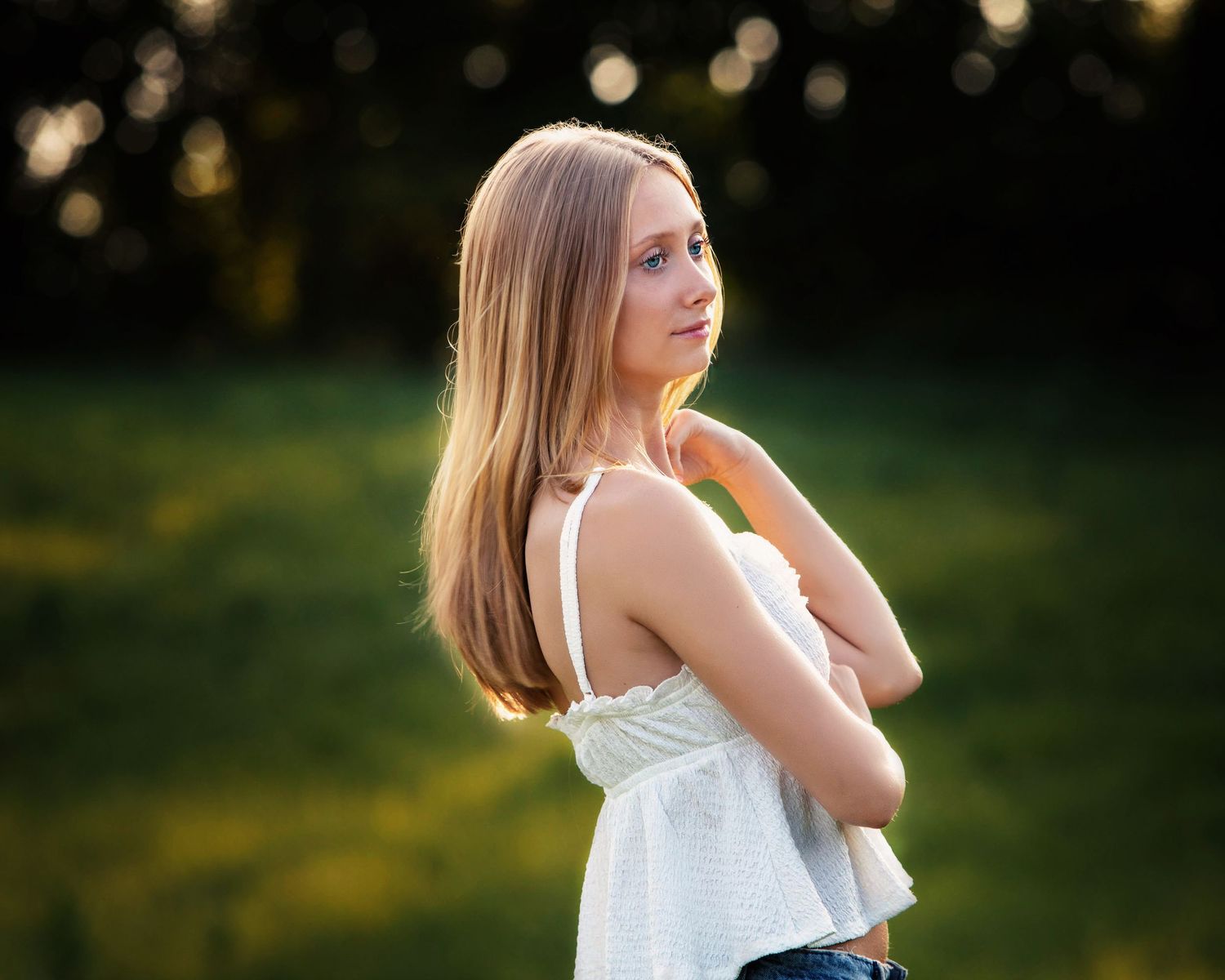 Young woman with long blonde hair in white top standing outdoors in golden hour sunlight with green bokeh background.