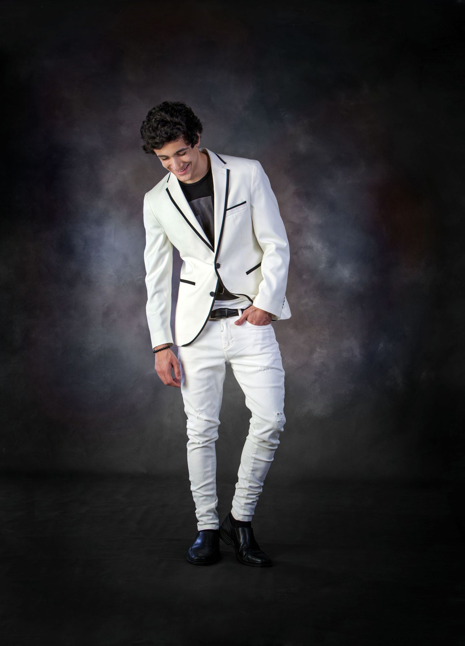Young man poses in white blazer and white jeans against dark studio backdrop.