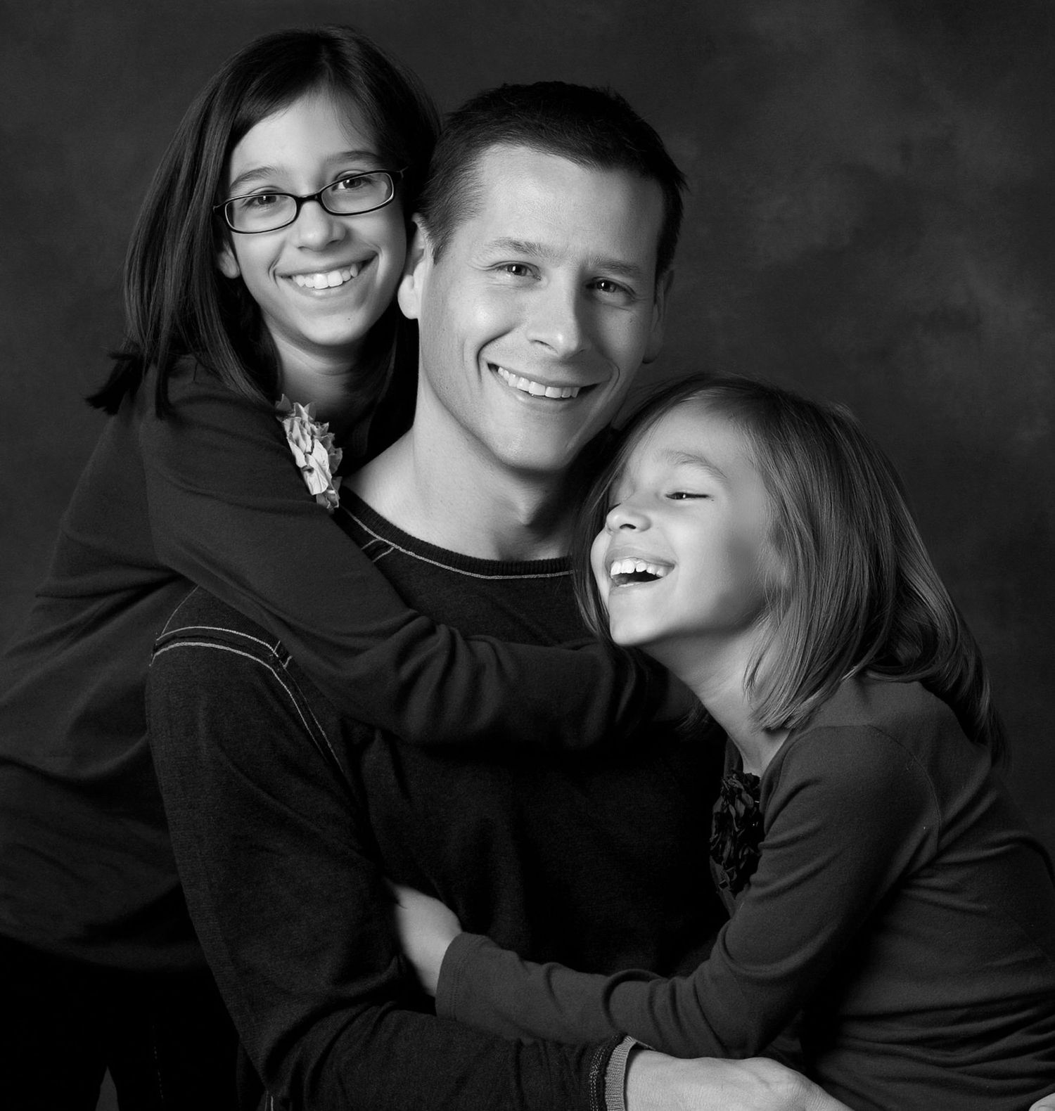 Black and white portrait of a smiling man hugging two laughing girls in a warm family studio photo.