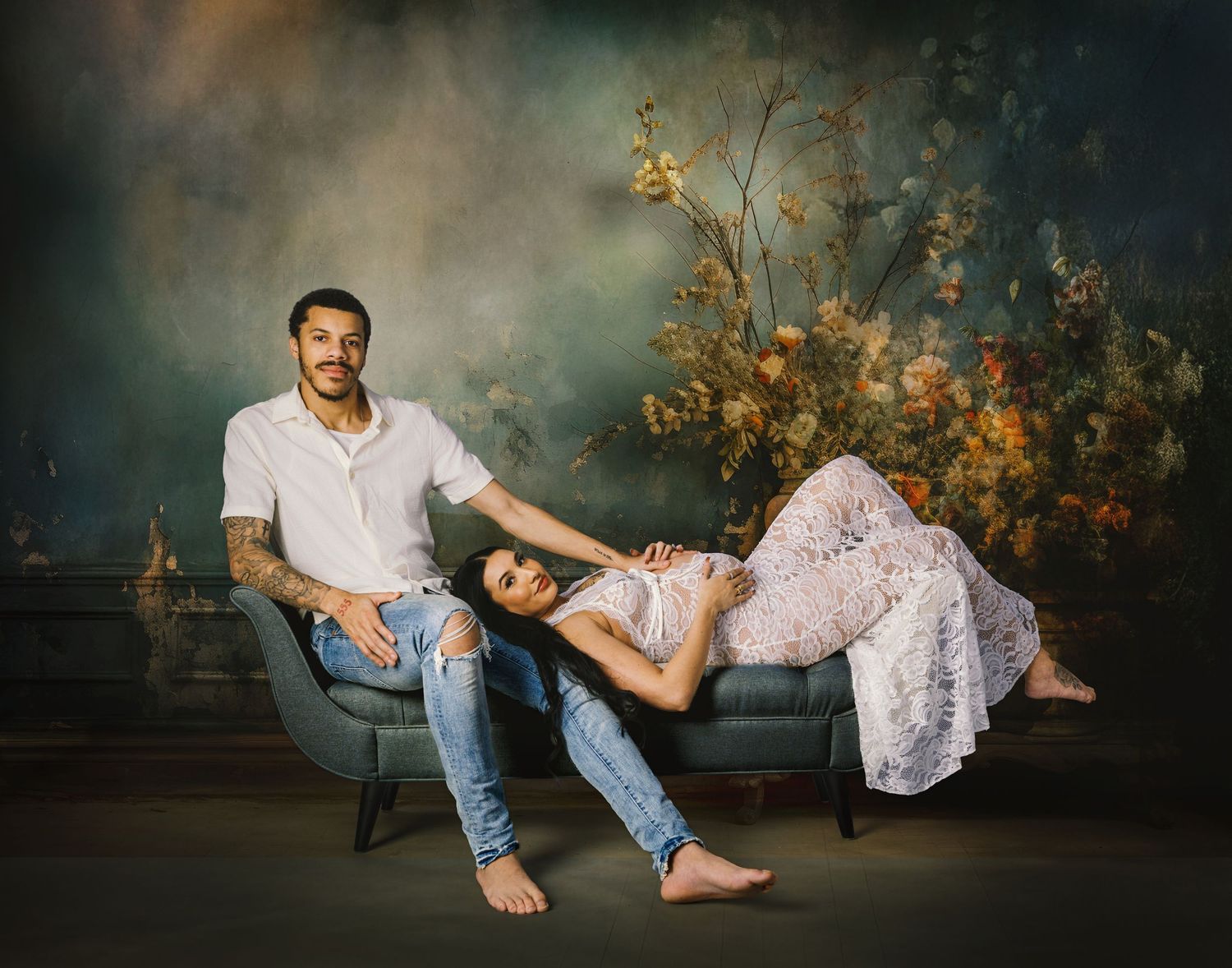 Couple posing for maternity photo shoot on teal sofa with floral backdrop, woman in white lace dress reclining.