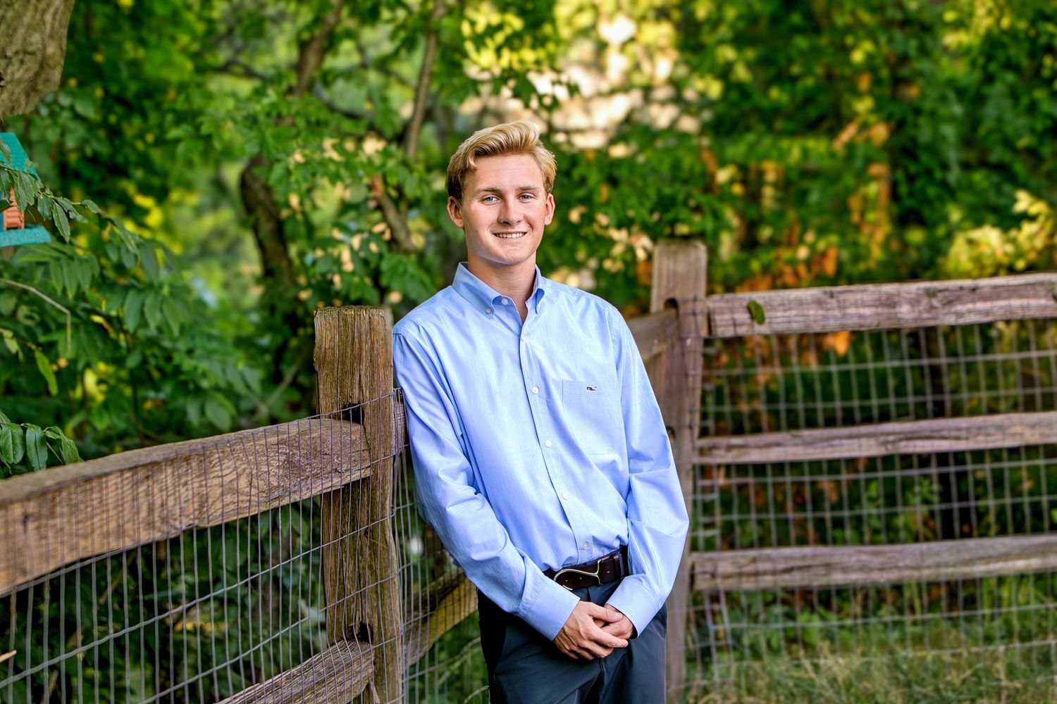Smiling young man in blue button-down shirt standing by wooden fence surrounded by green trees.