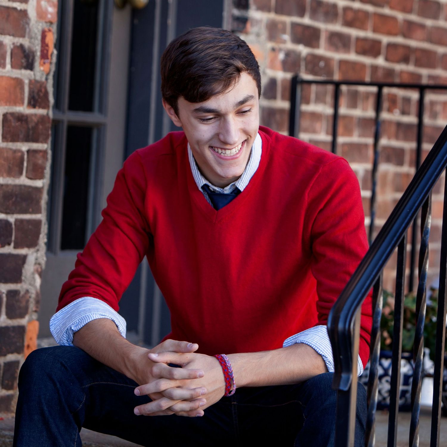Senior boy in red sweater and tie smiling while sitting on brick steps outdoors, by Jocelyn M. Photography in Rochester, MI