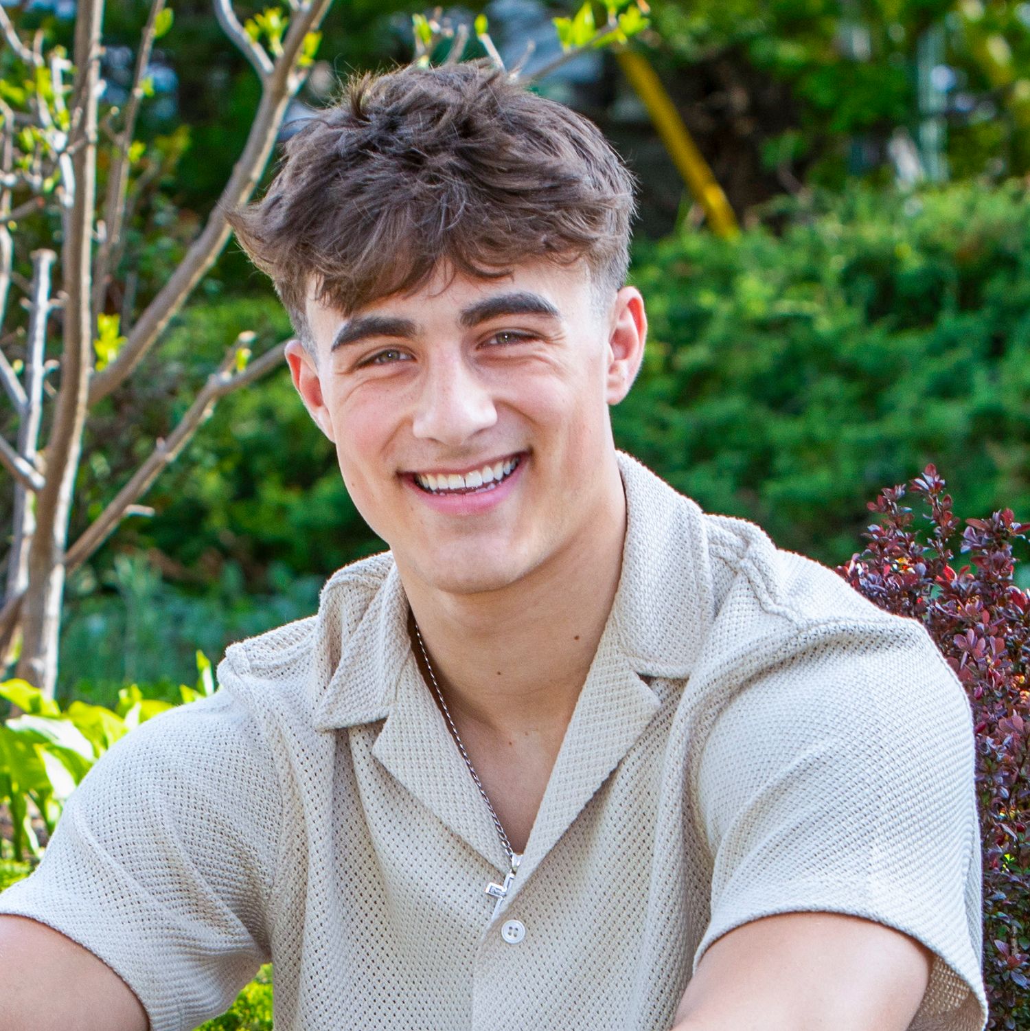 Young man with brown hair smiling outdoors, wearing a light beige polo shirt with green trees in background.