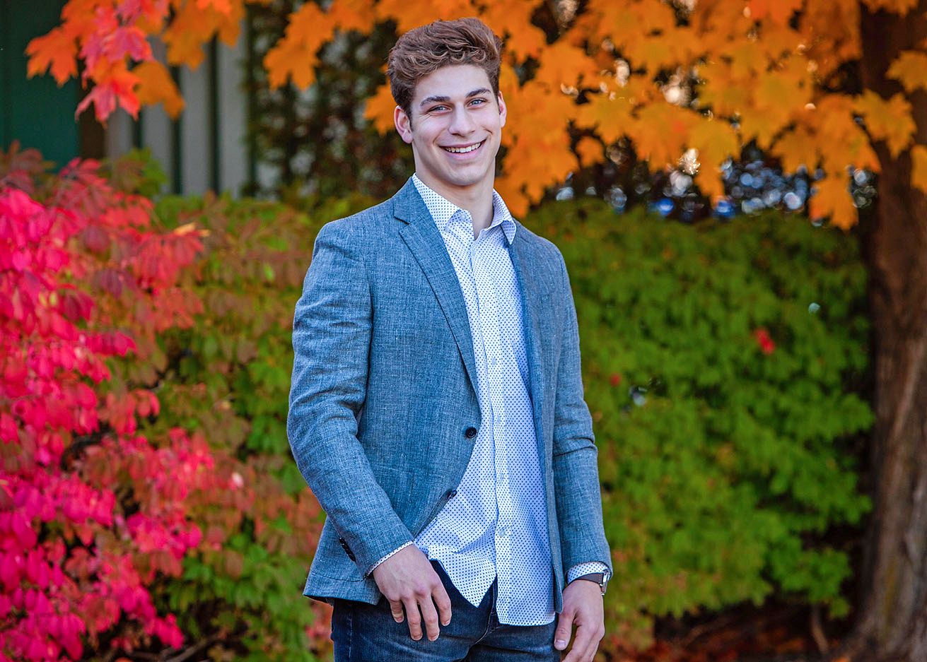 Smiling young man in gray blazer posing outdoors among vibrant red and orange autumn leaves.