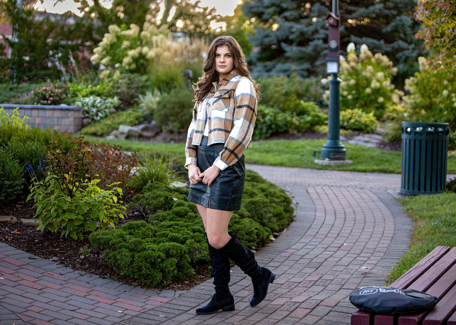 Young woman in plaid top and leather skirt with knee-high boots poses in garden.