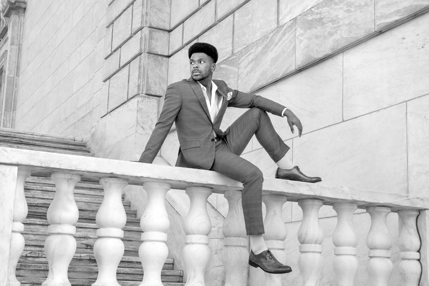 A stylish man in a tailored suit and beret poses on ornate white stone balustrade steps in a black and white photo.