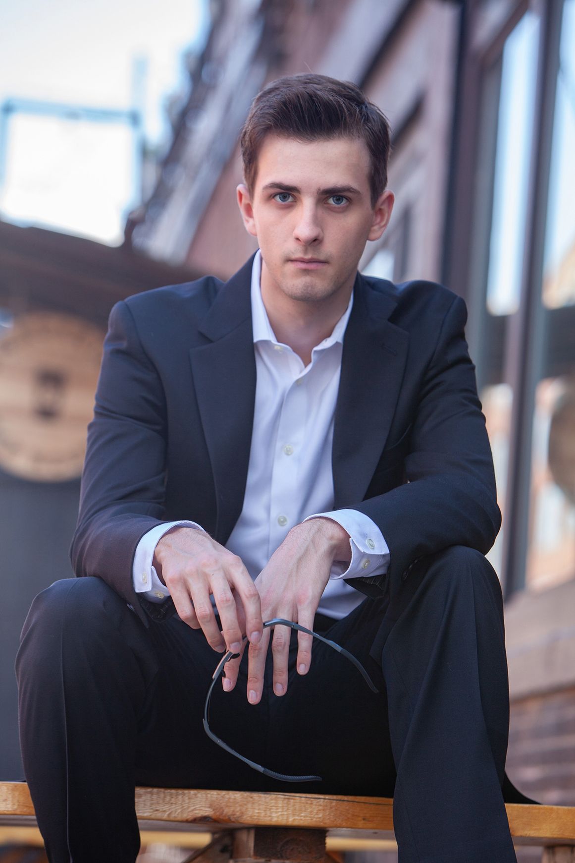 Elegant man in black suit sitting outdoors holding sunglasses in urban setting.