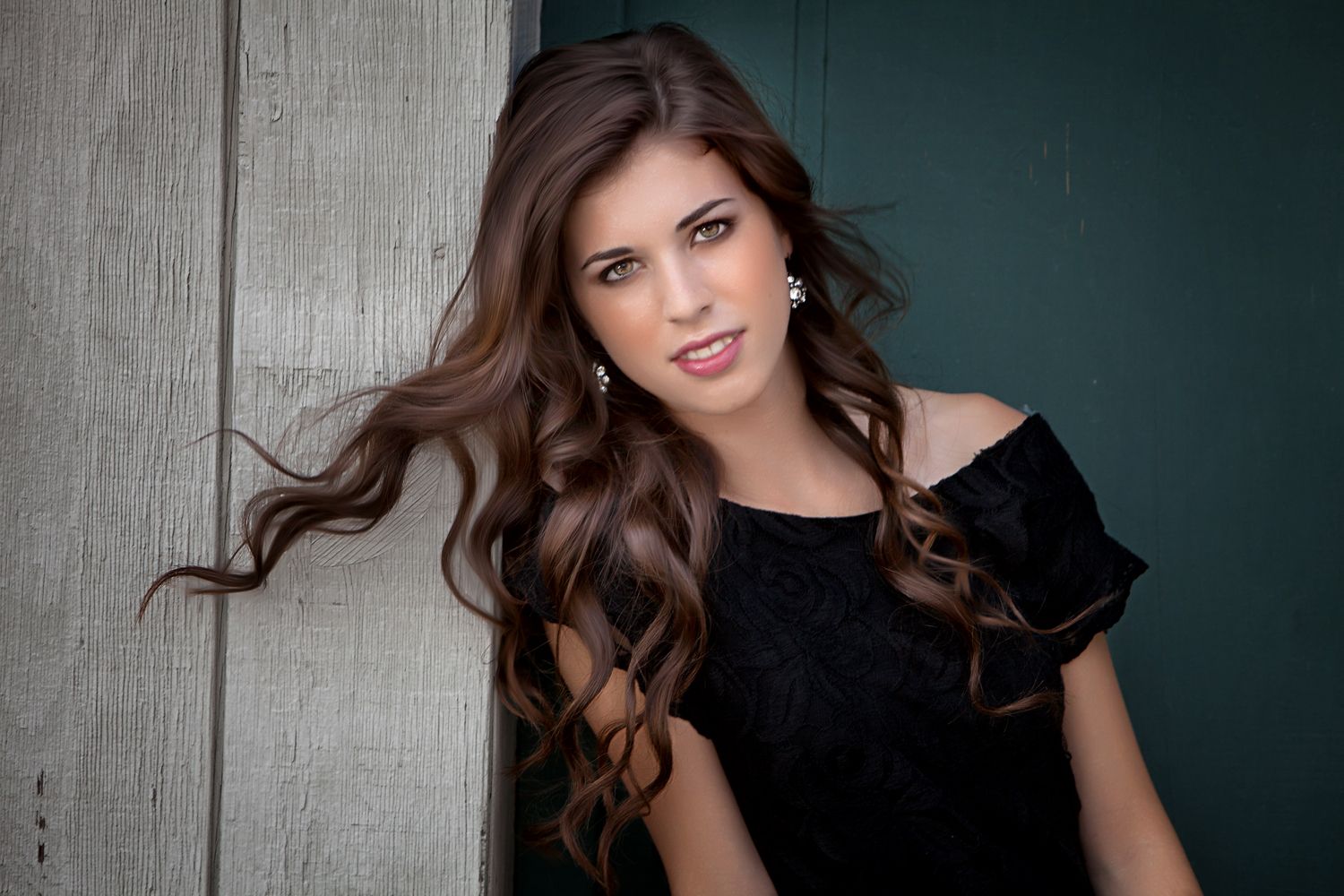 Young woman with long brunette hair in black off-shoulder top poses against rustic wooden wall.