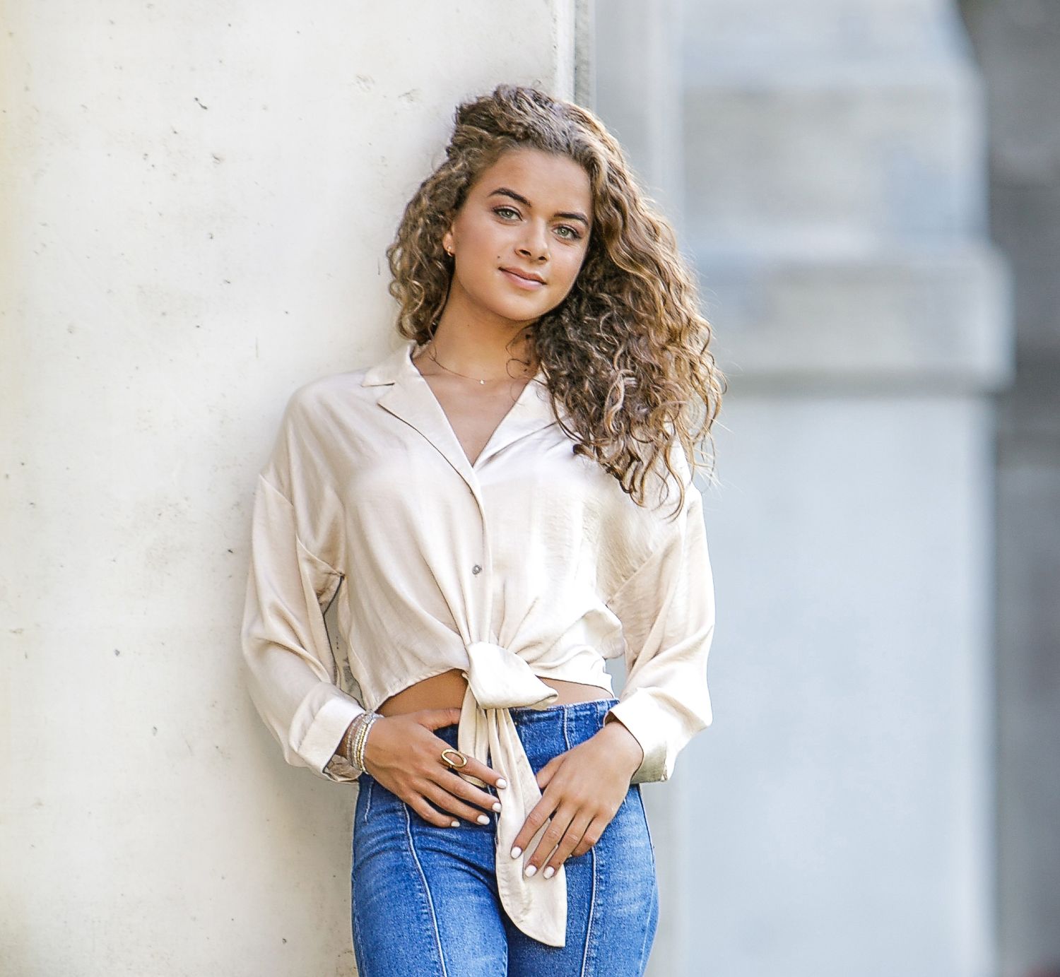 Young woman with curly hair wearing a tied white shirt and blue jeans, leaning against a wall outdoors.