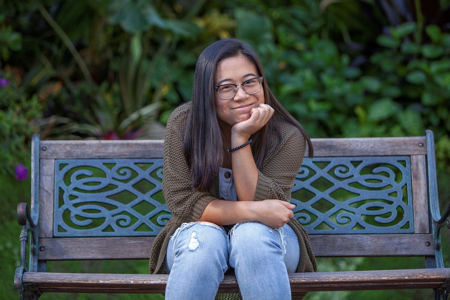 Smiling young woman with glasses sitting on an ornate garden bench surrounded by lush green plants.