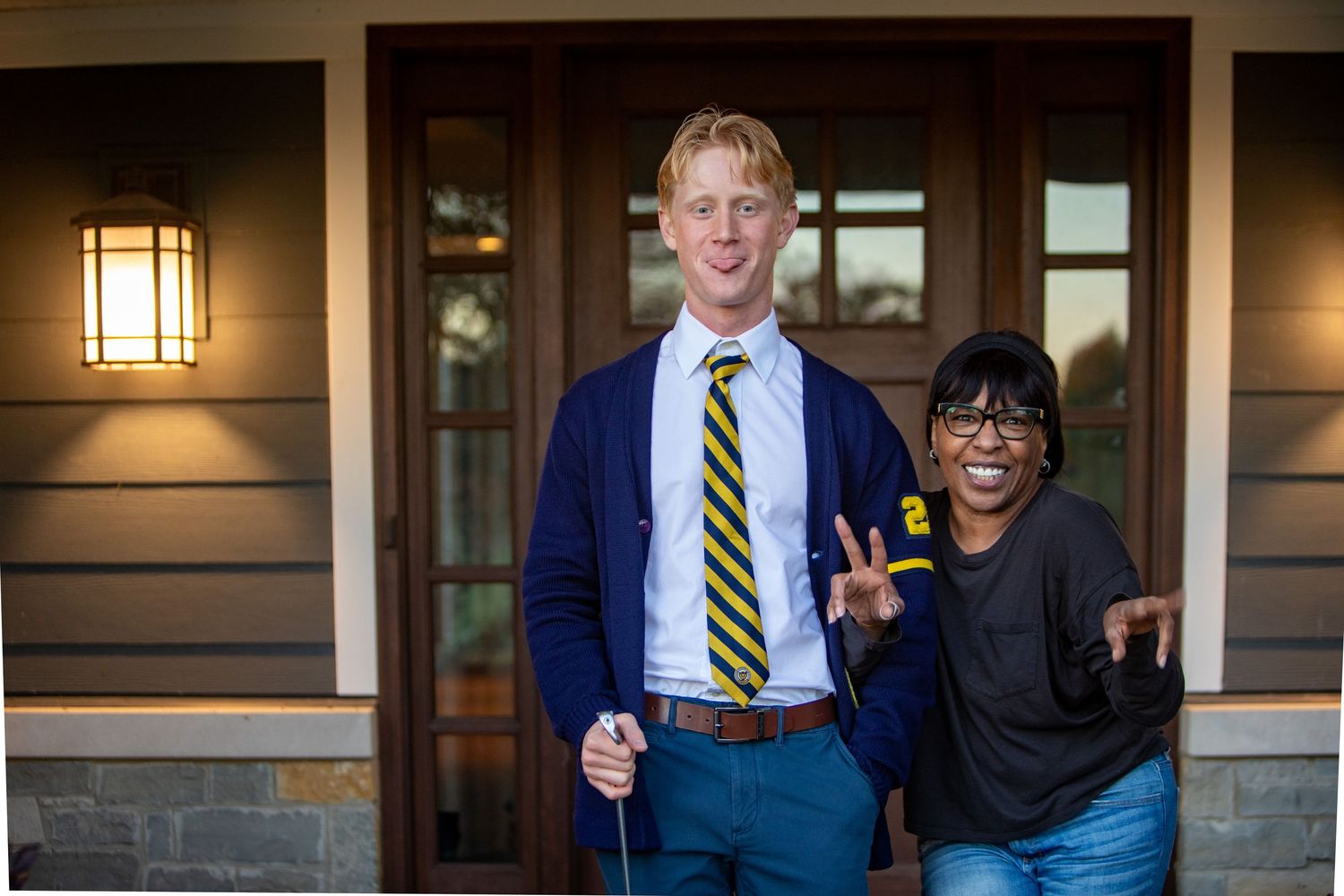 Tall young man in navy cardigan and striped tie poses with a woman outside a modern home.