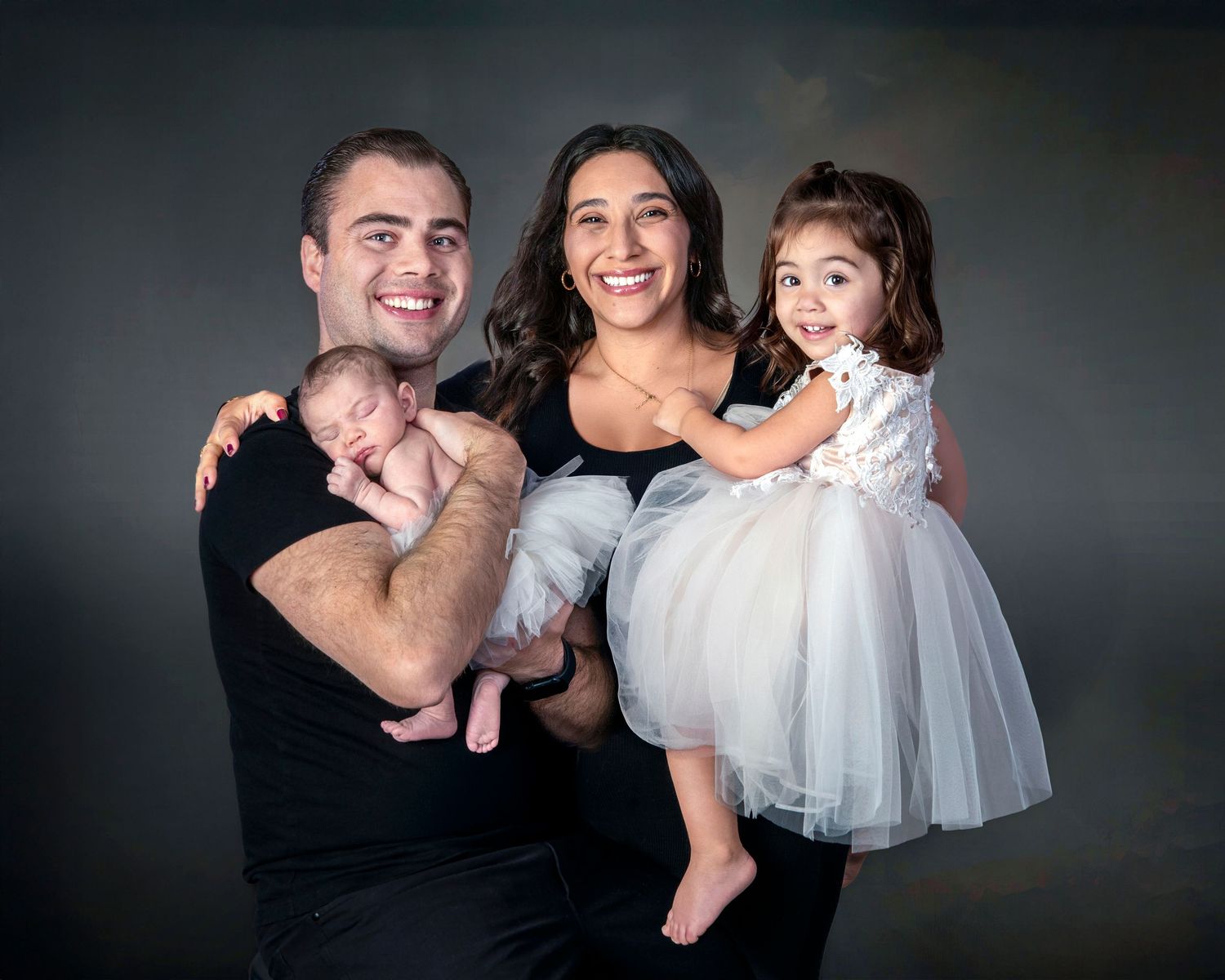 Happy family portrait with parents holding newborn baby and toddler daughter in white tutu dress against gray backdrop.