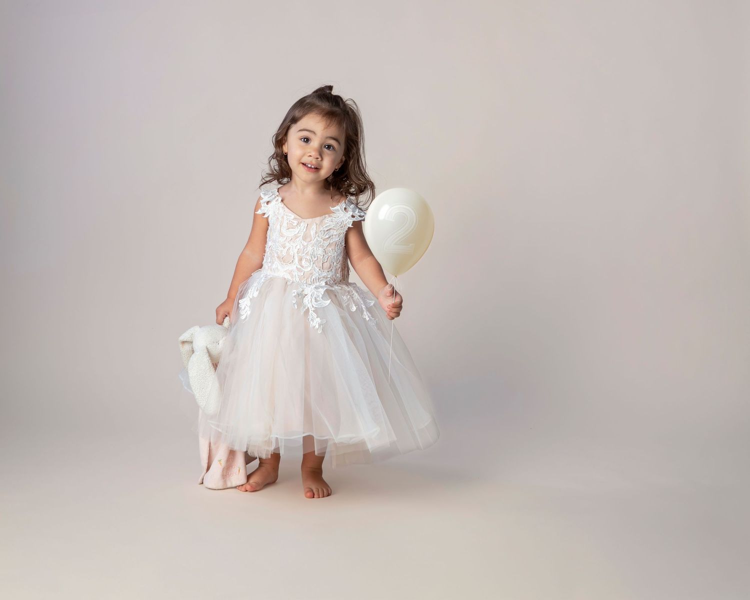 Young girl in white lace tutu dress holding balloon and toy lamb, posing barefoot in studio photo.