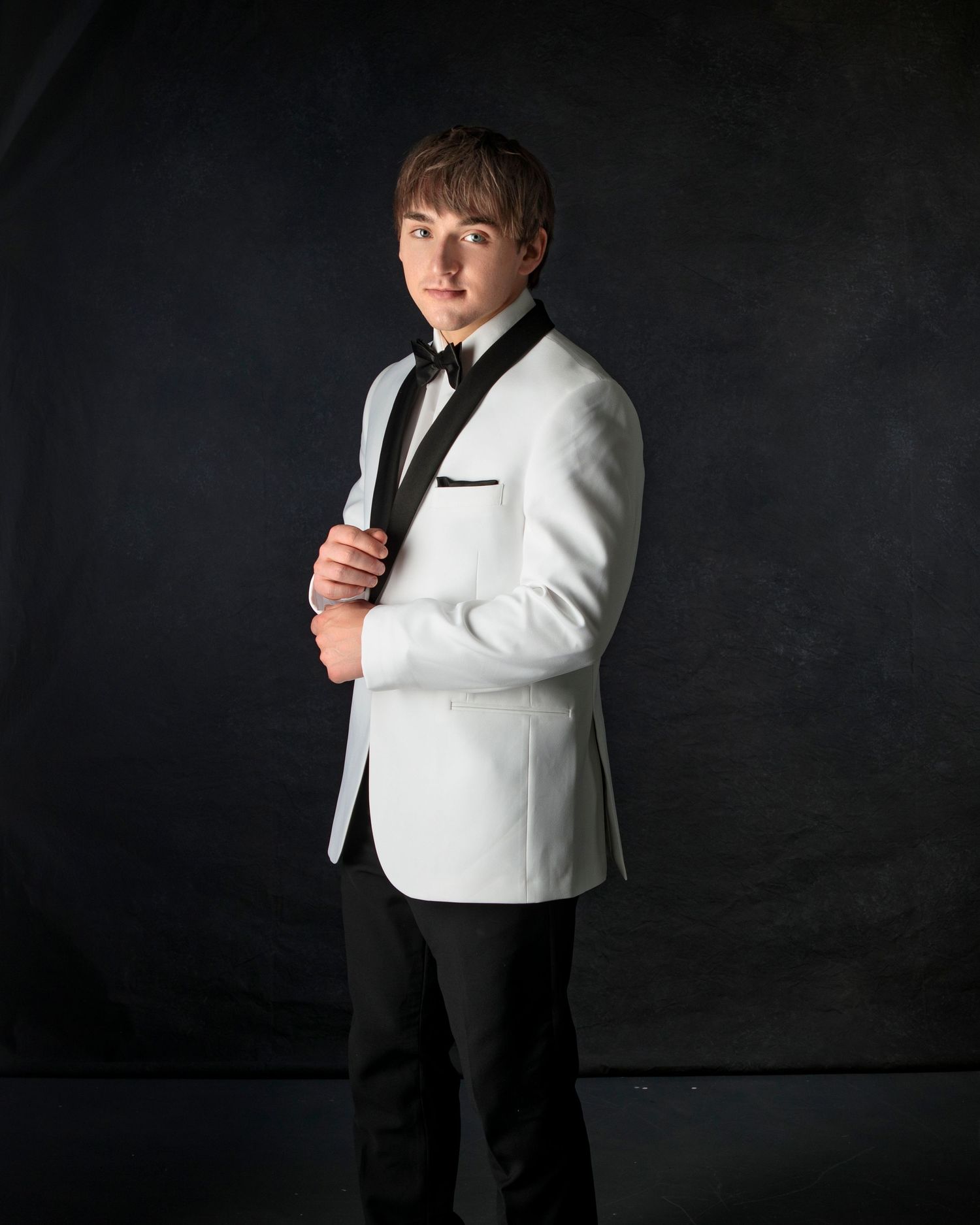 Young man in white tuxedo jacket with black lapels and bow tie poses against dark background.