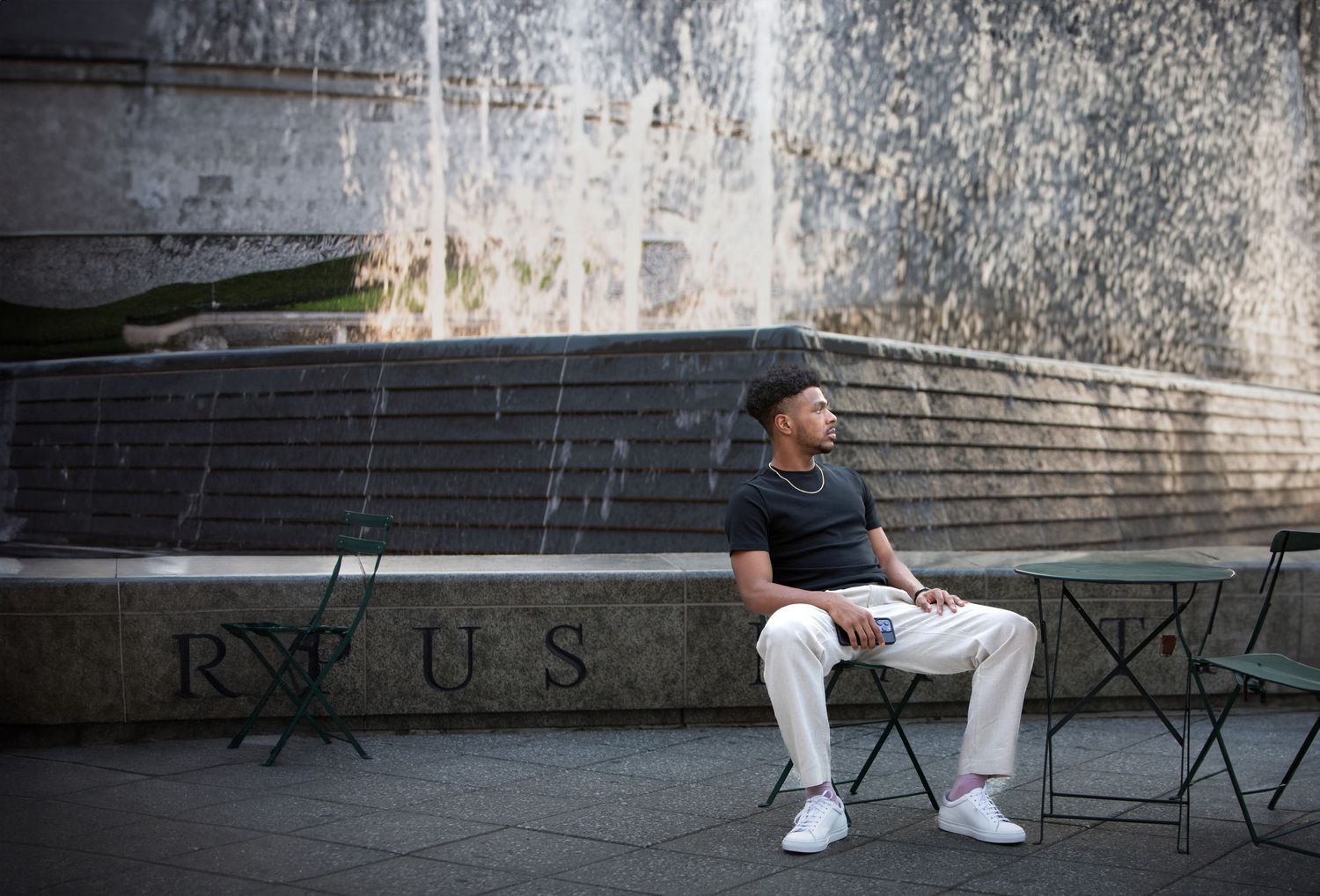 Young man in white pants and dark shirt sits on bench near decorative fountain outdoors.