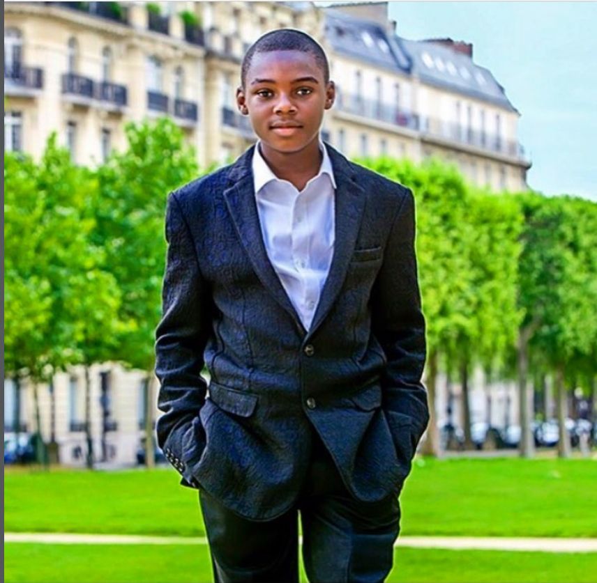 Young man in a dark suit and white shirt standing on a green lawn with Parisian architecture in the background.
