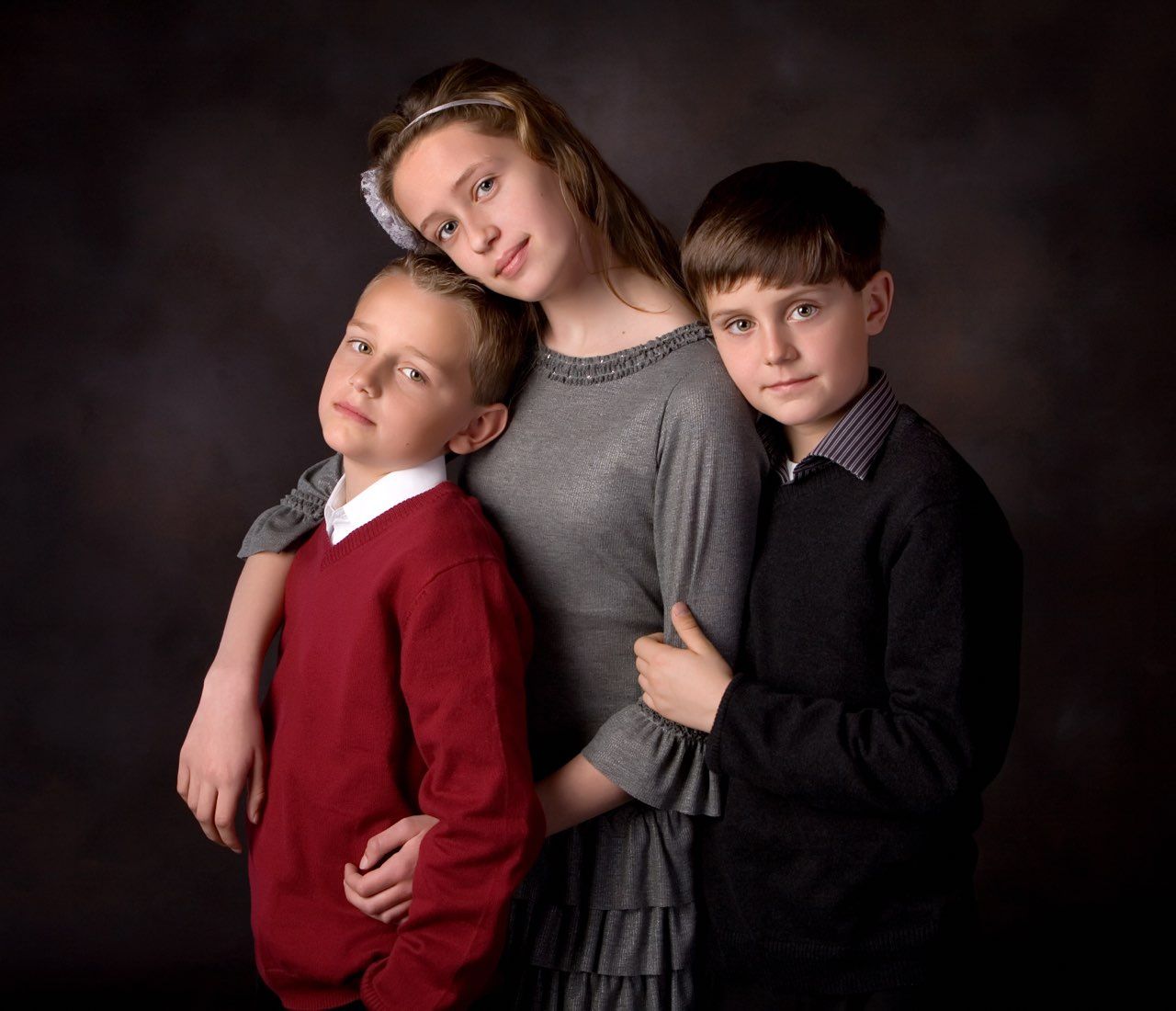 Three children pose for a formal portrait, two boys in red and black and a girl in gray, against a dark studio background.