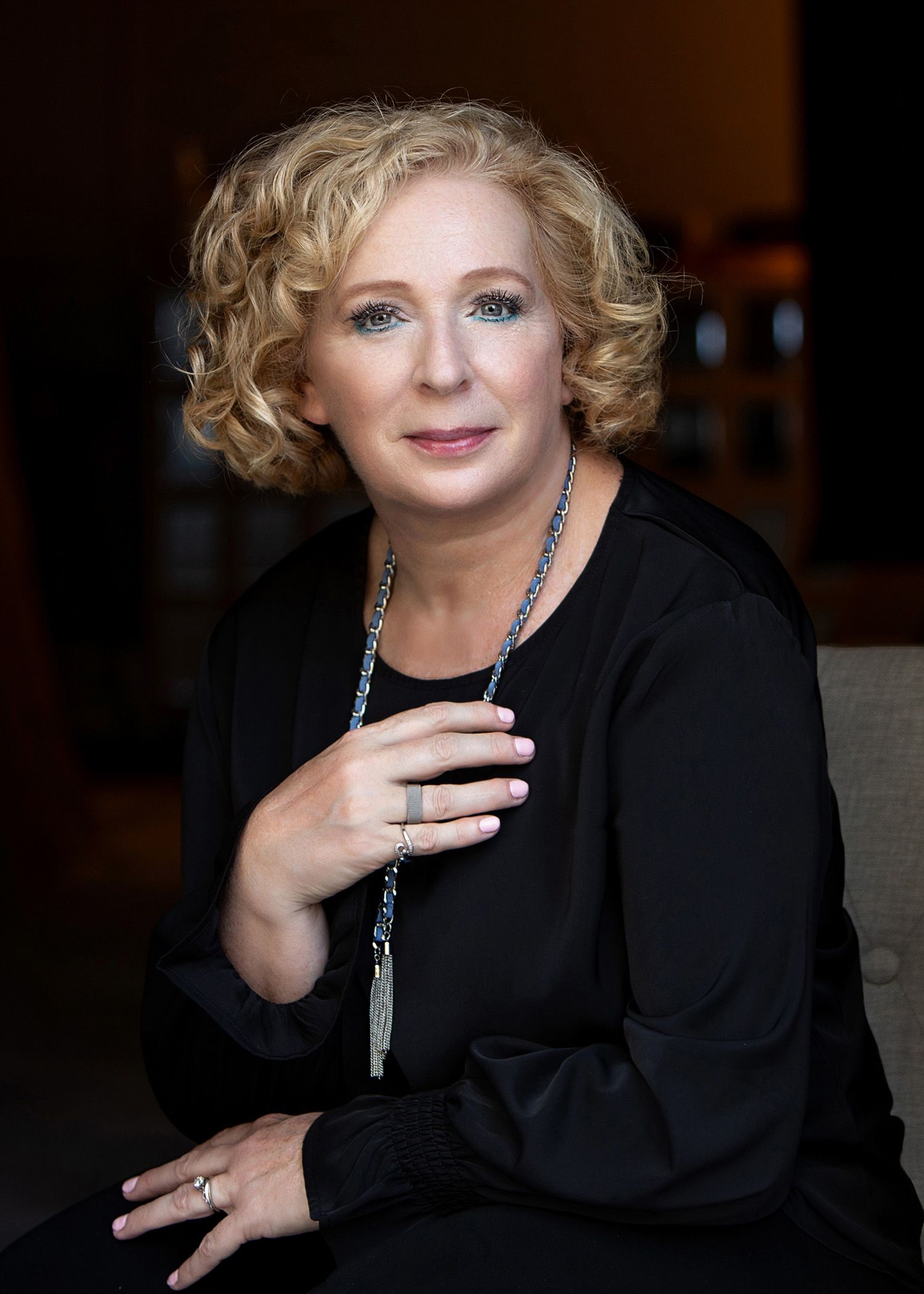 Sophisticated woman with curly blonde hair in black outfit with beaded necklace in warm setting.