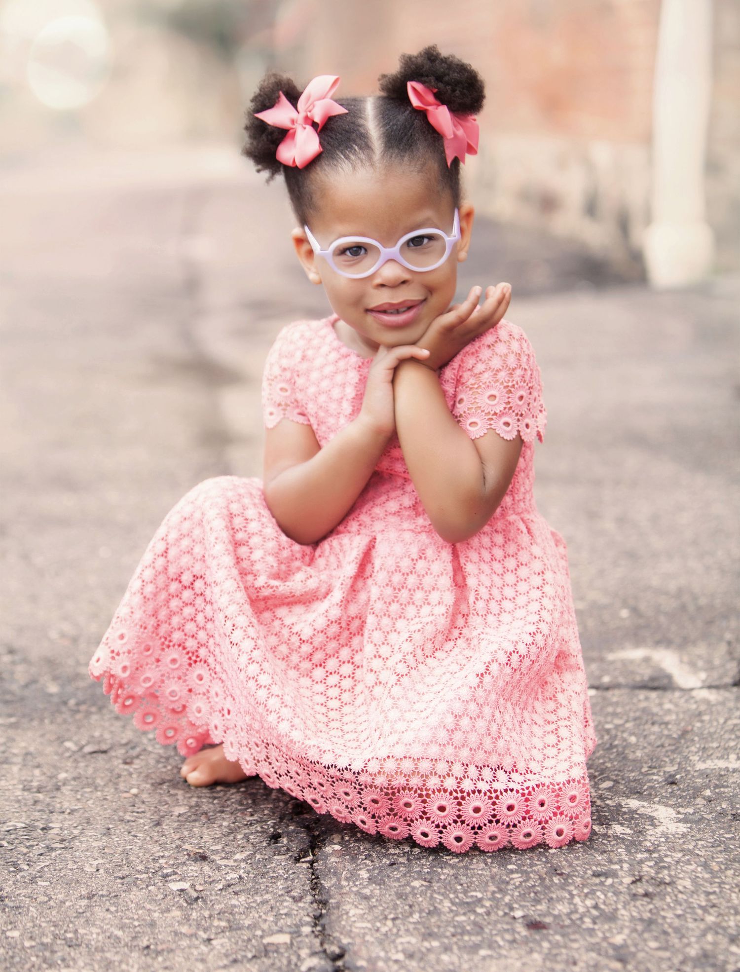Adorable little girl in pink eyelet dress and glasses posing on outdoor pavement.