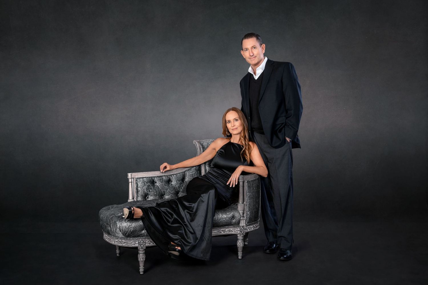 Elegant couple in black formal attire posing with a silver tufted chaise lounge against a dark studio background.