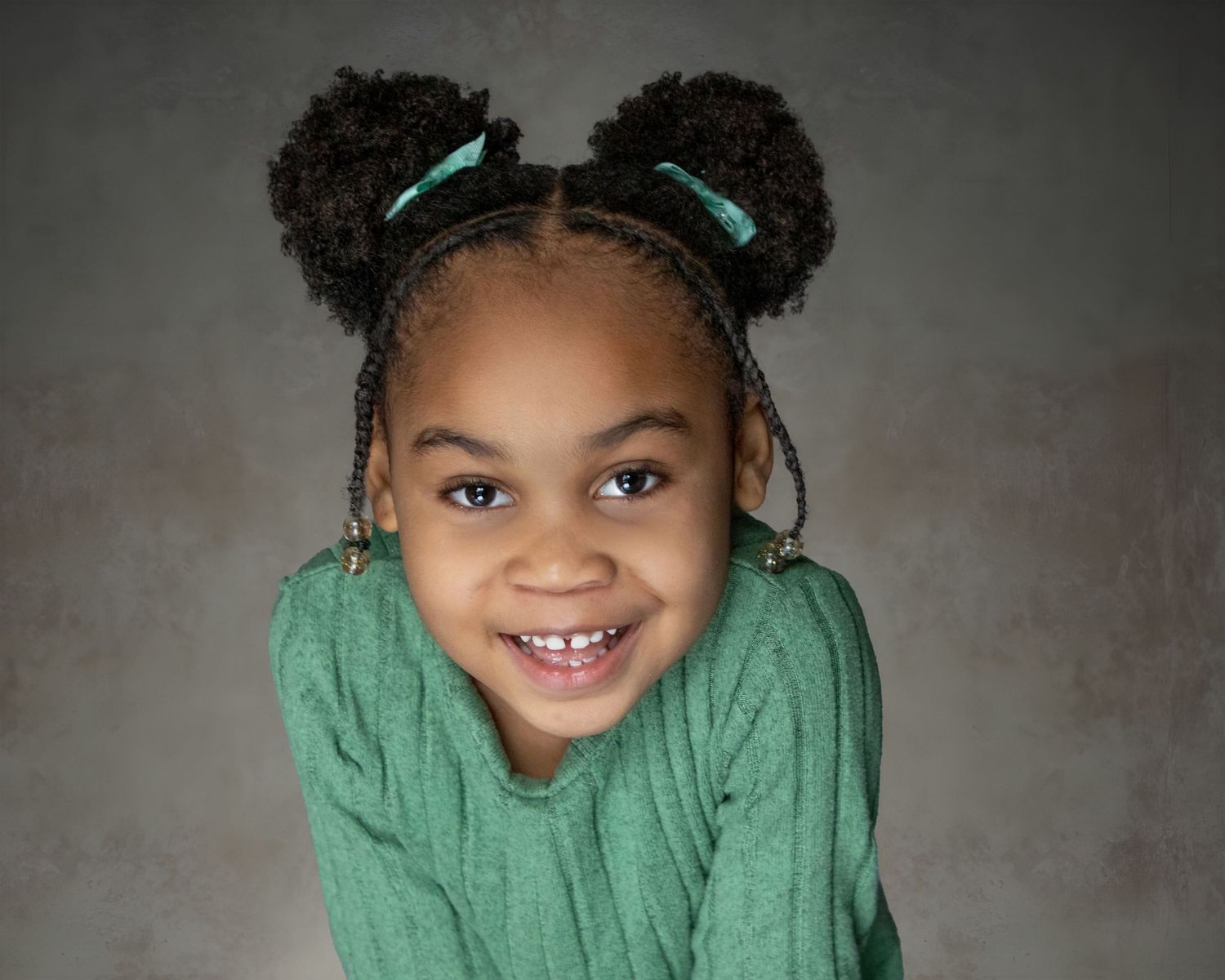 Adorable young girl with braided buns and green ribbons smiling brightly in a green sweater.
