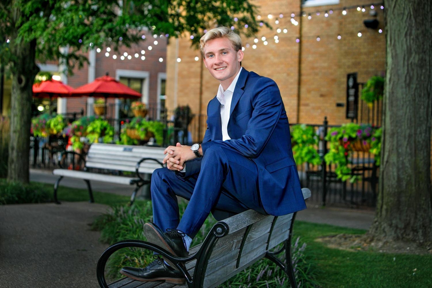 Young man in blue suit sitting on bench in outdoor garden setting with string lights and greenery.