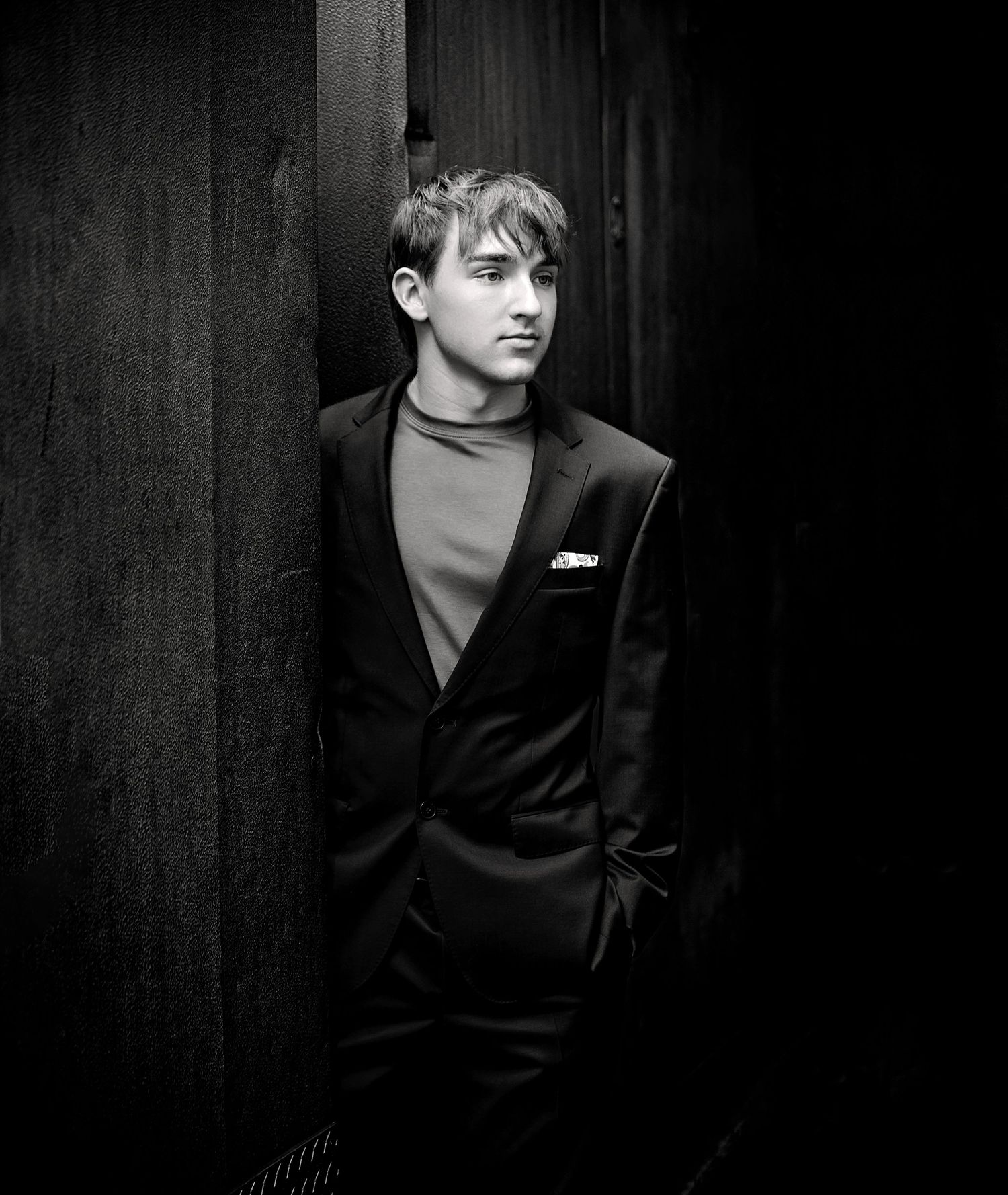 Black and white portrait of a young man in a suit leaning against a dark wooden wall, looking away thoughtfully.