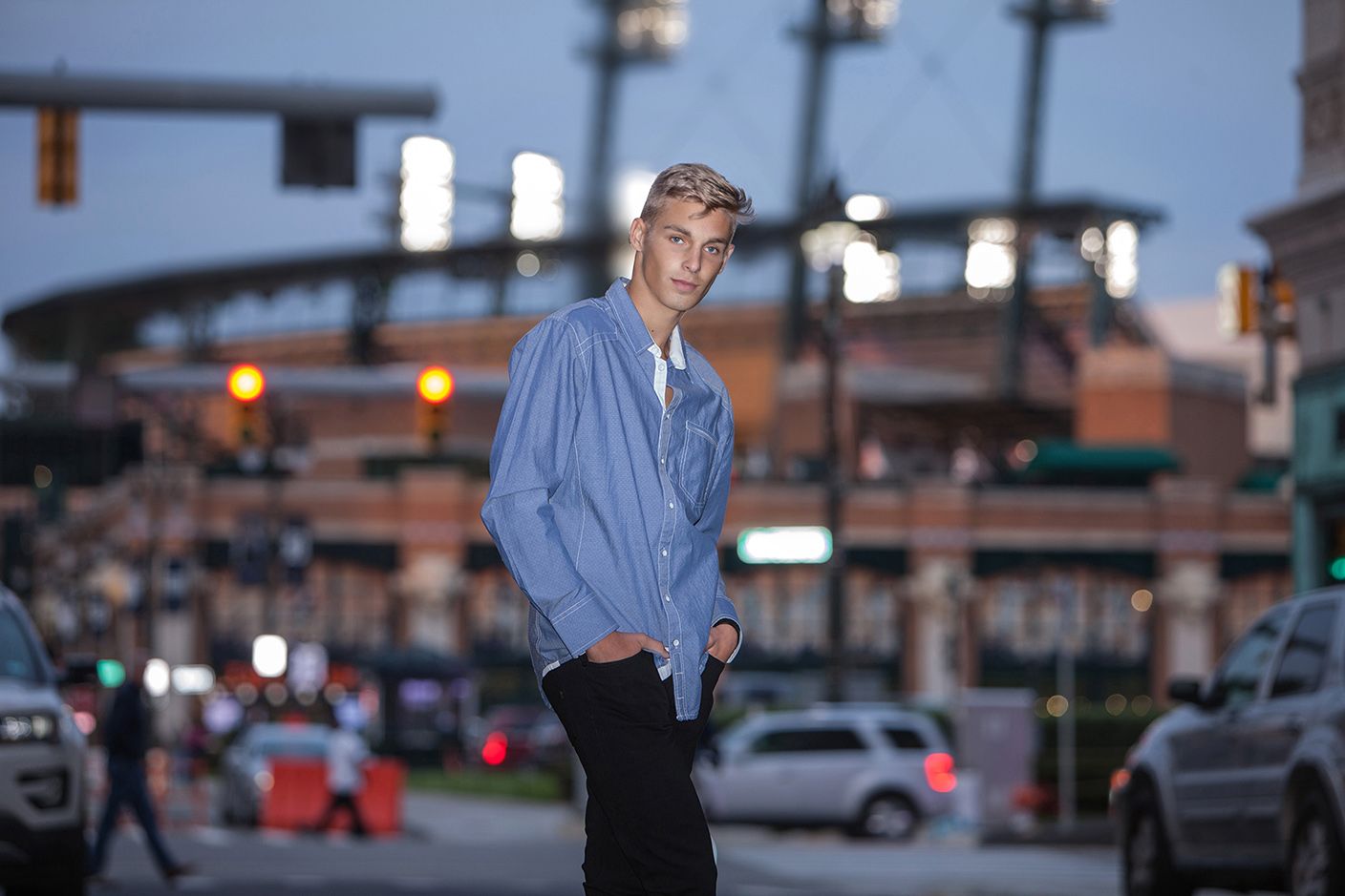 Young man in denim shirt standing casually at city intersection with stadium lights at dusk.