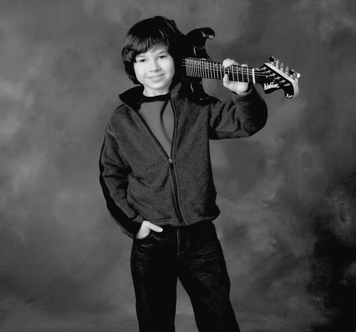 Smiling boy poses with electric guitar on shoulder in black and white studio portrait photo.