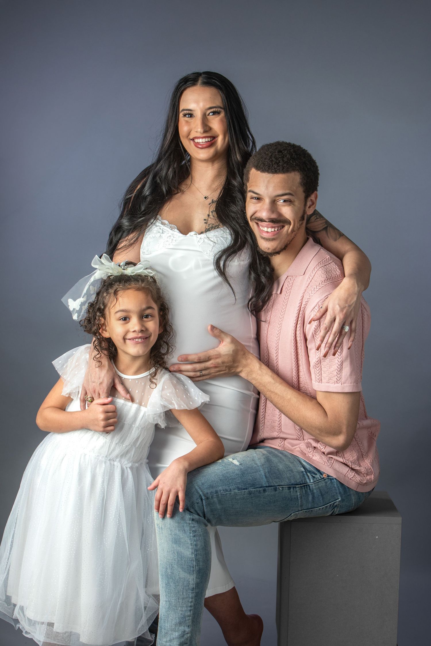A smiling pregnant woman in white dress poses with her husband in pink shirt and young daughter in white dress for a maternity photo.