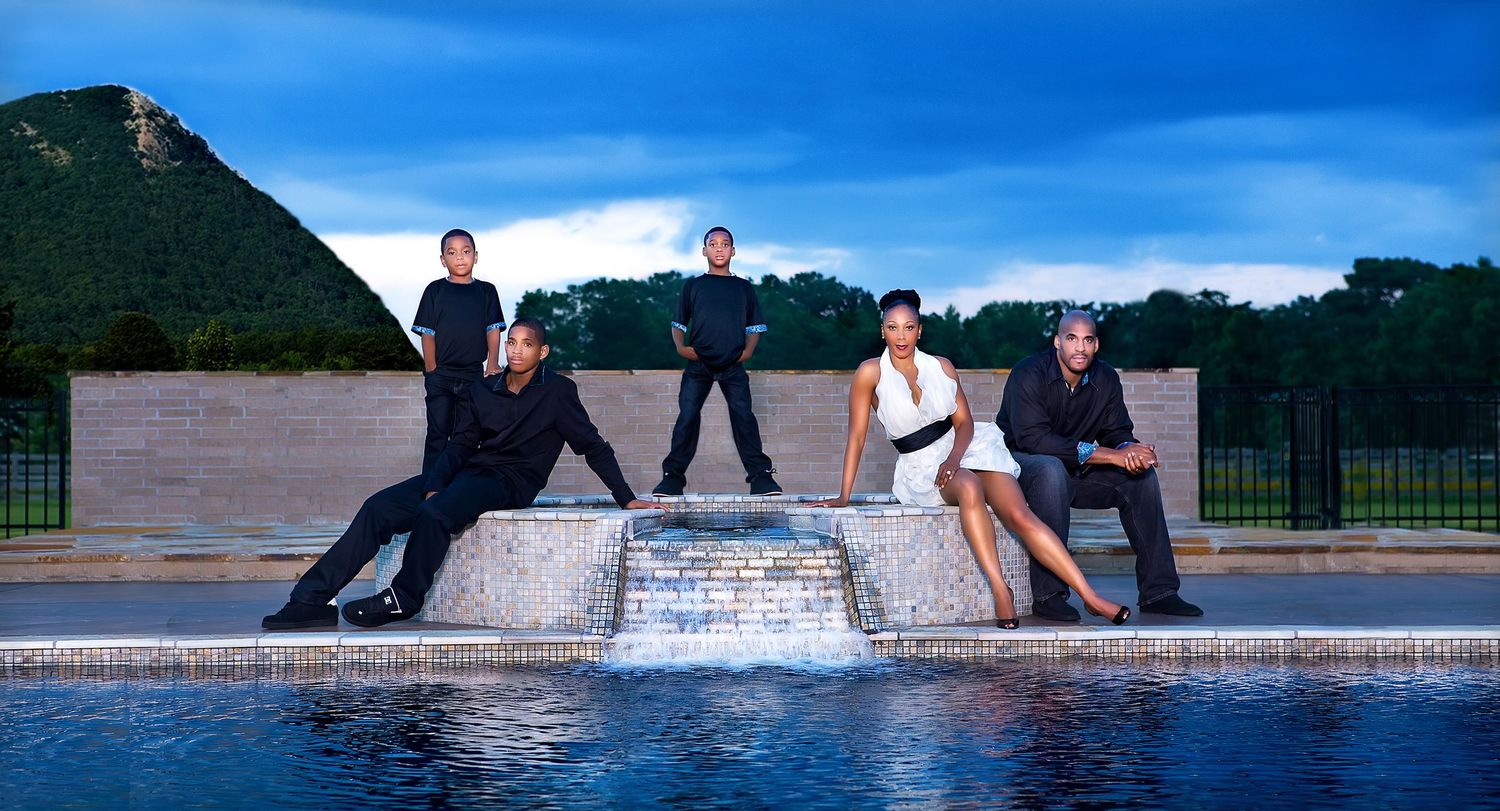 Group of five people posing elegantly by a decorative fountain pool at dusk with lush green hills in the background.