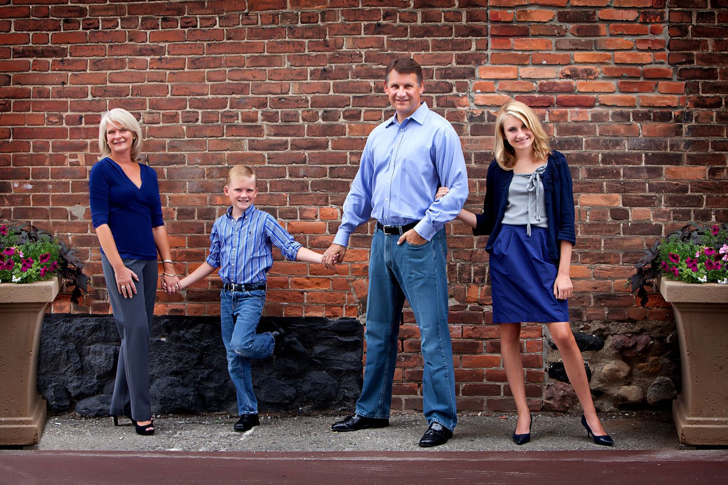 Family of four posing in front of a brick wall, parents holding hands with young boy in coordinated blue outfits.