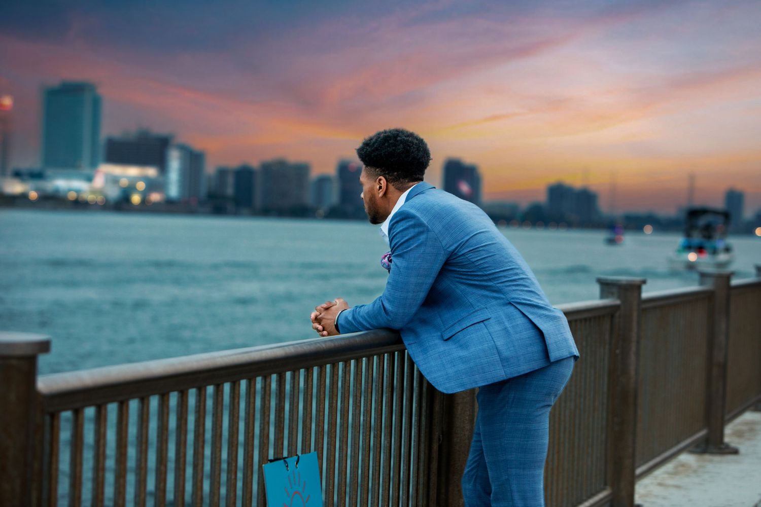 Man in blue plaid suit leaning on waterfront railing at sunset with city skyline in background.