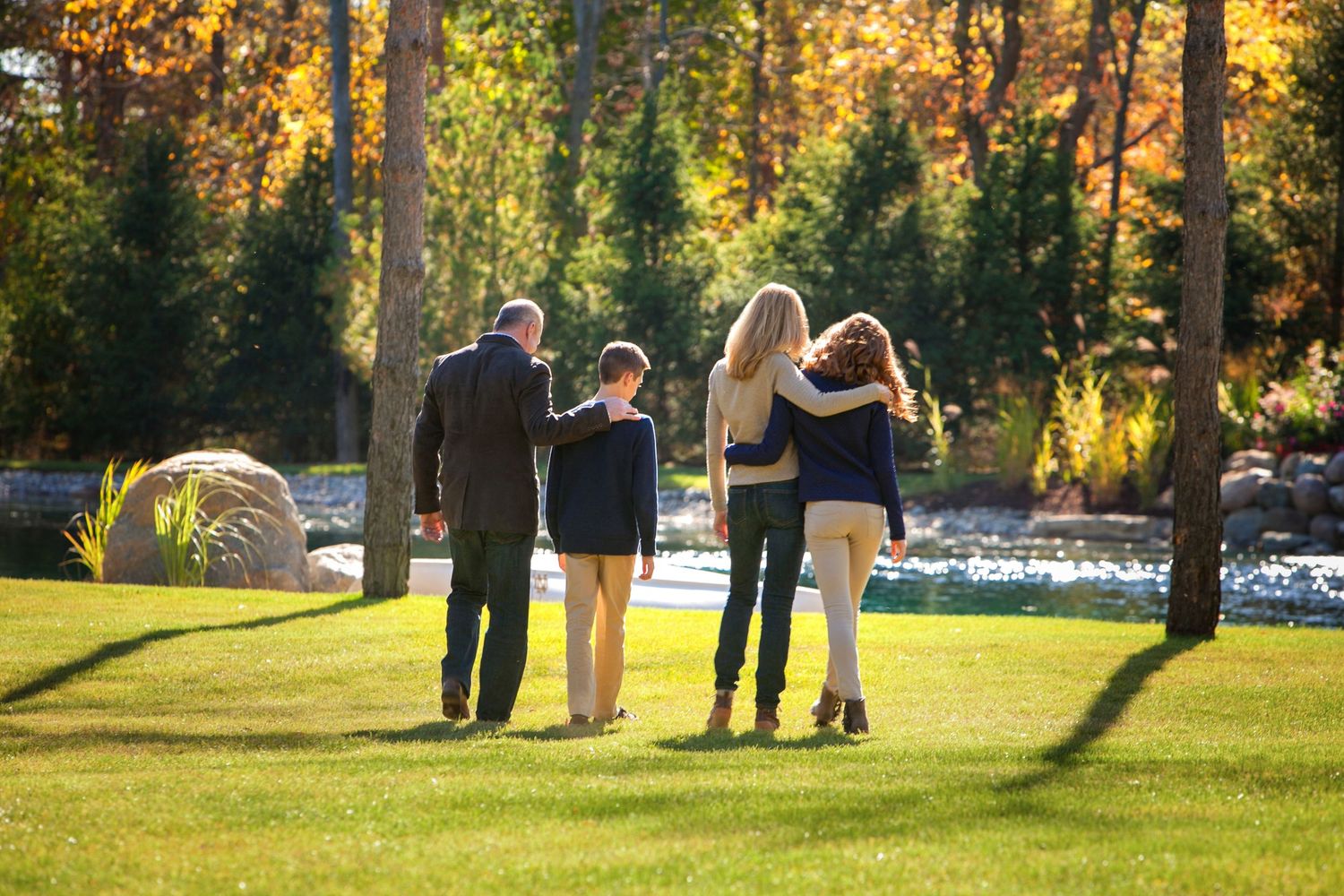 Family of four walking together through a lush green park with colorful autumn trees in the background.