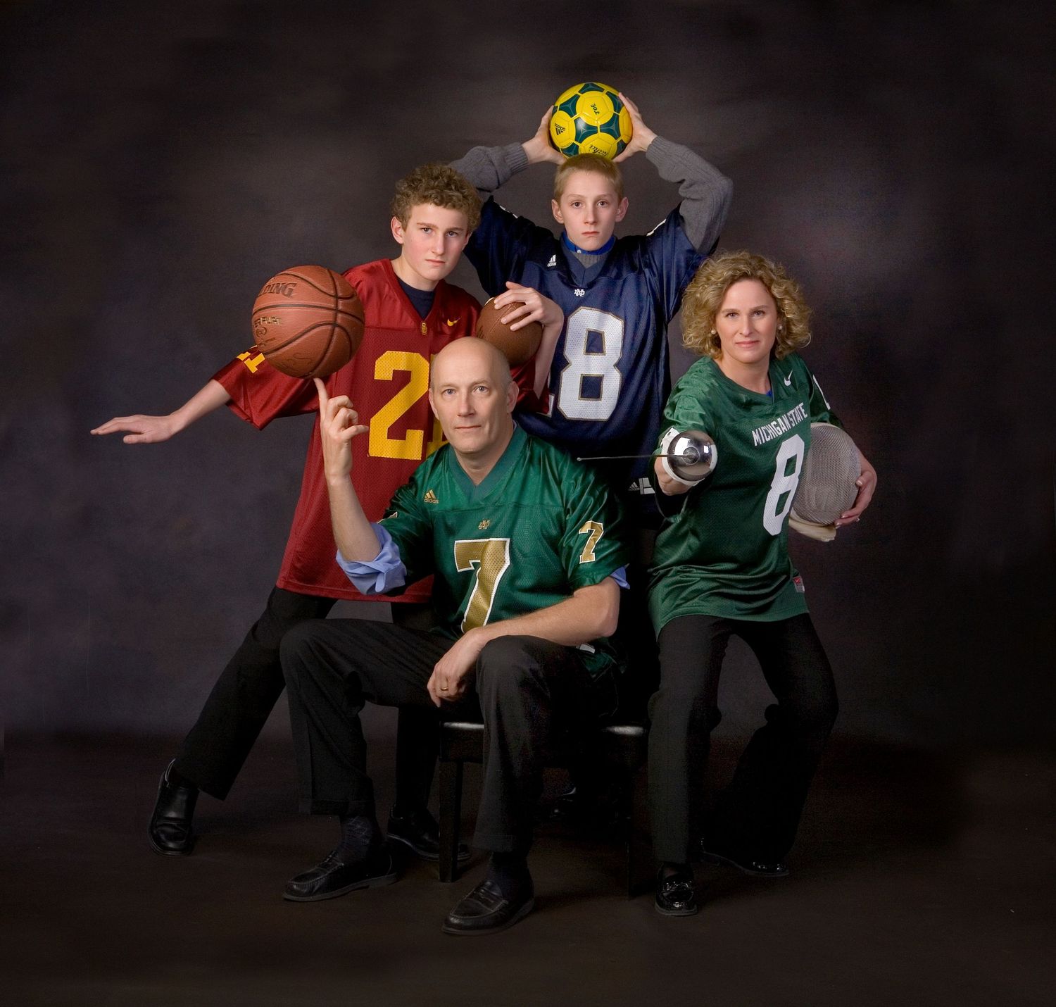 Family of four posing in sports jerseys with basketball, soccer ball, and football for a studio portrait photo.
