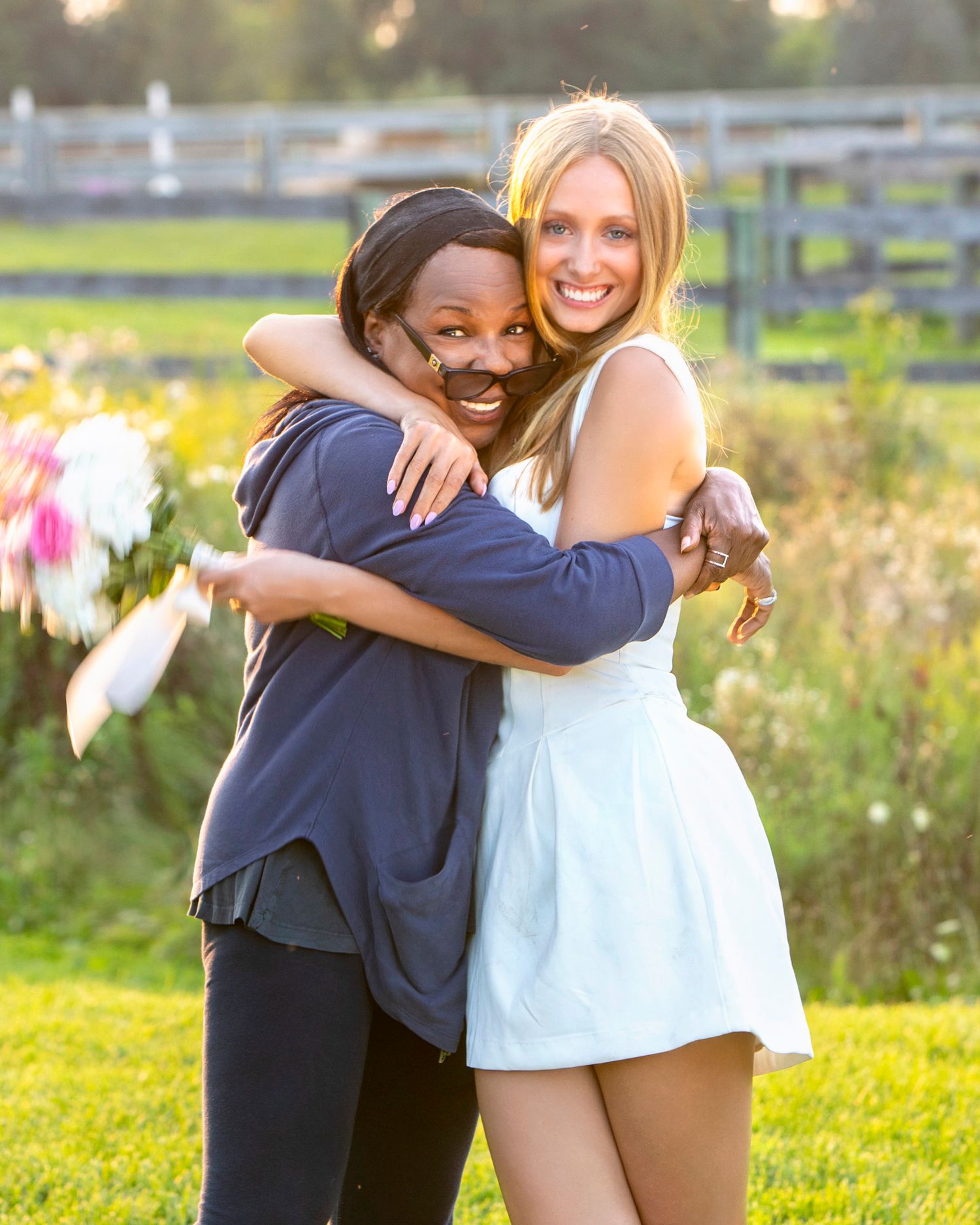 Two women sharing a warm hug outdoors on a sunny day in a green field setting.