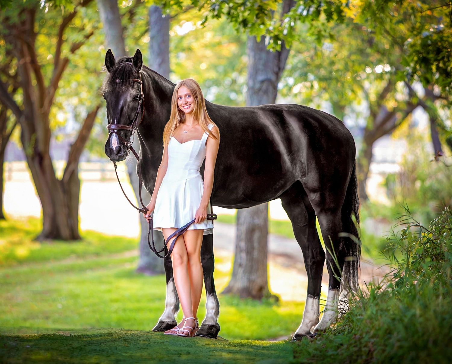 Young blonde woman in white dress posing with a black horse on green grass surrounded by trees.
