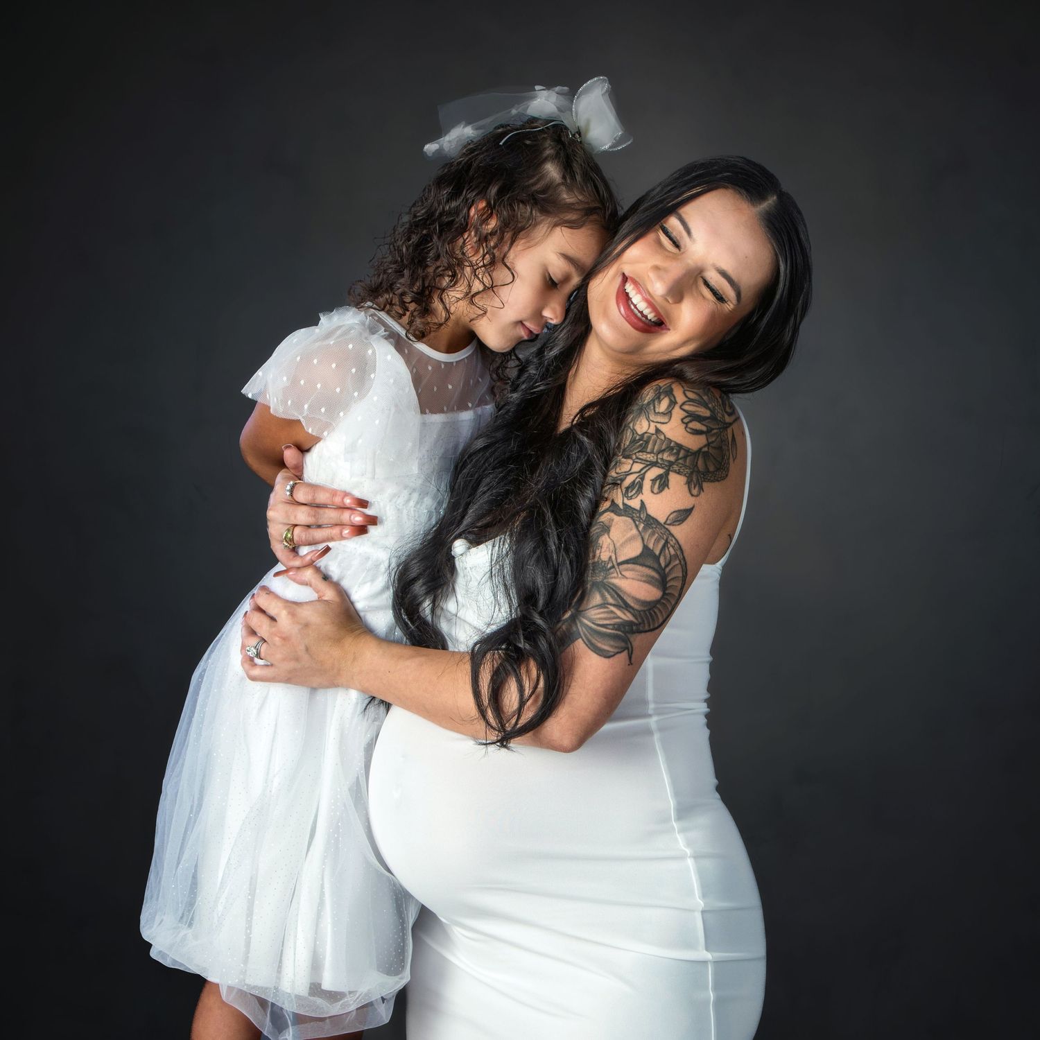Pregnant woman in white dress hugs smiling young girl in white dress during maternity photo session.