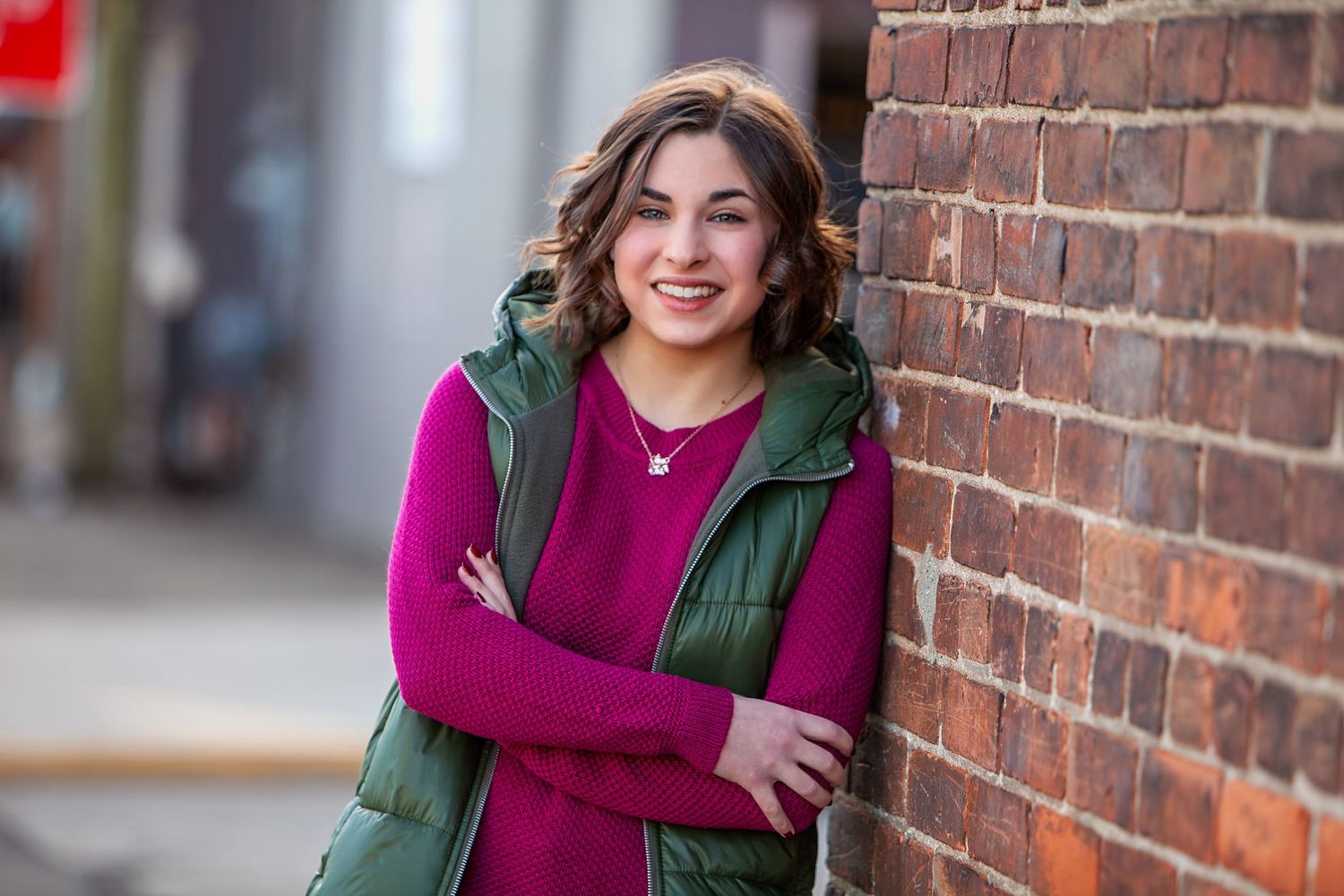 Senior girl in green vest and pink top leaning against brick wall outdoors, by Jocelyn M. Photography in Rochester, MI