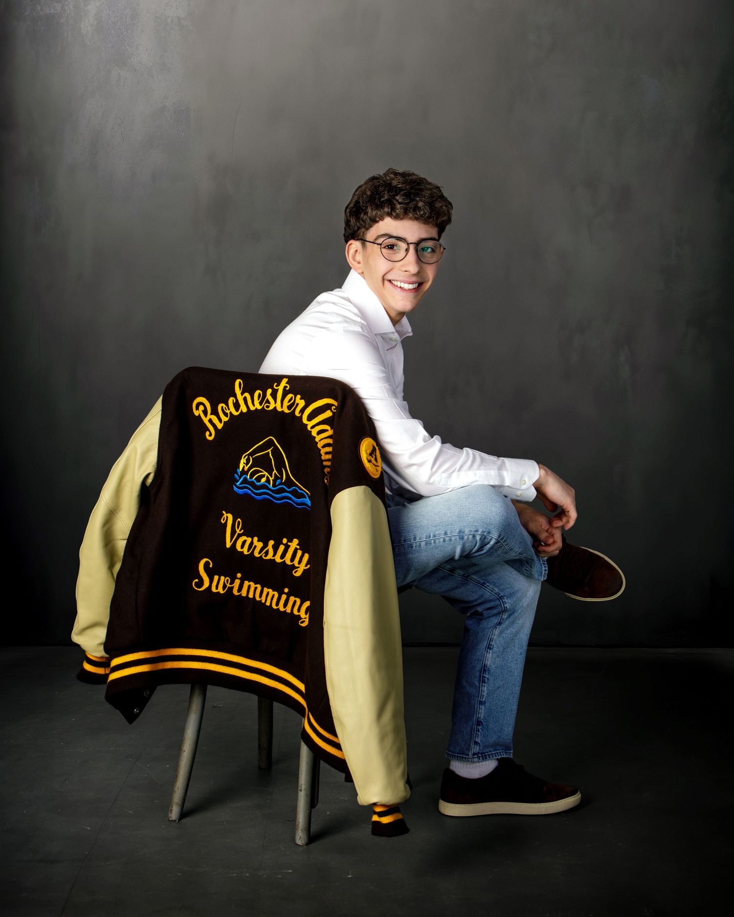 Smiling teen boy in glasses sitting with Rochester Clam Varsity Swimming letterman jacket for senior portrait.
