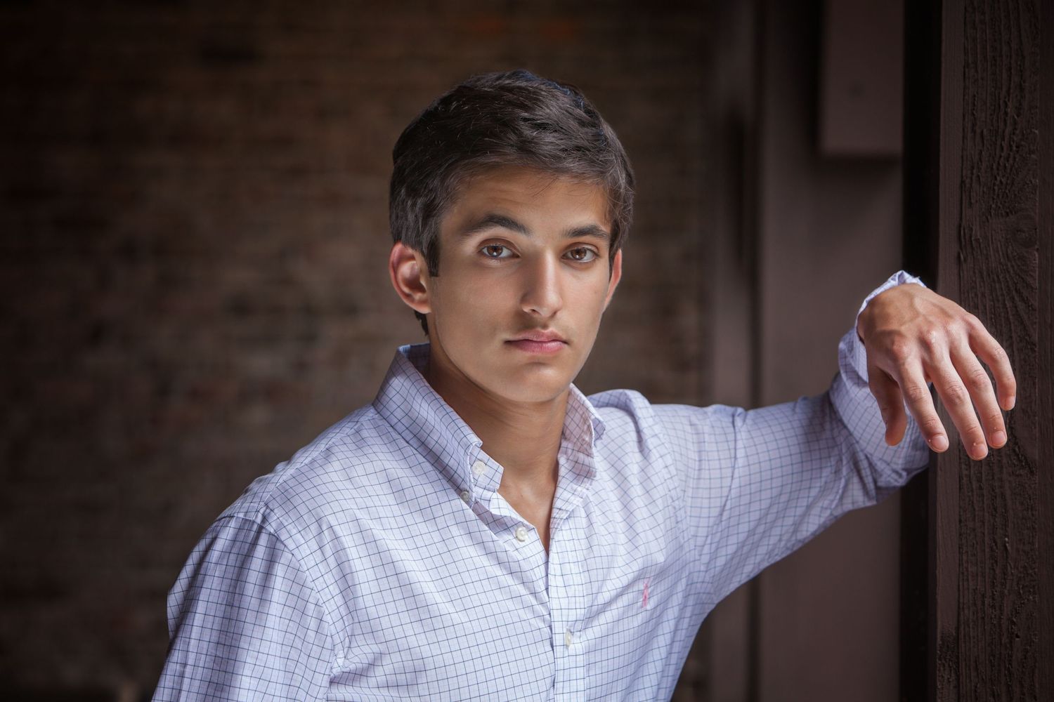 Young man in a light blue checkered shirt leaning against a wooden door frame with a brick wall background.