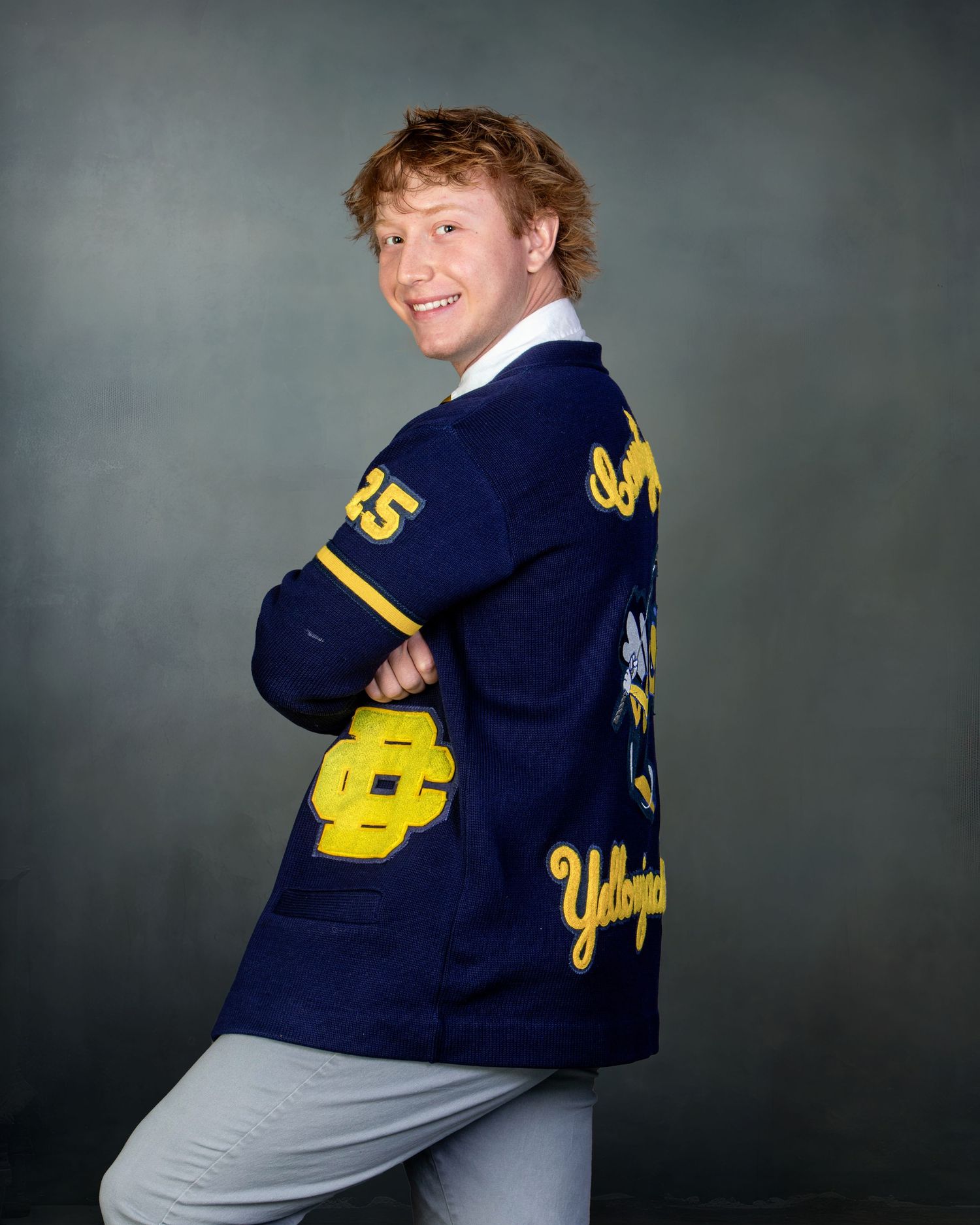 Smiling teen boy in navy yellow letterman jacket posing sideways for senior portrait against gray background.